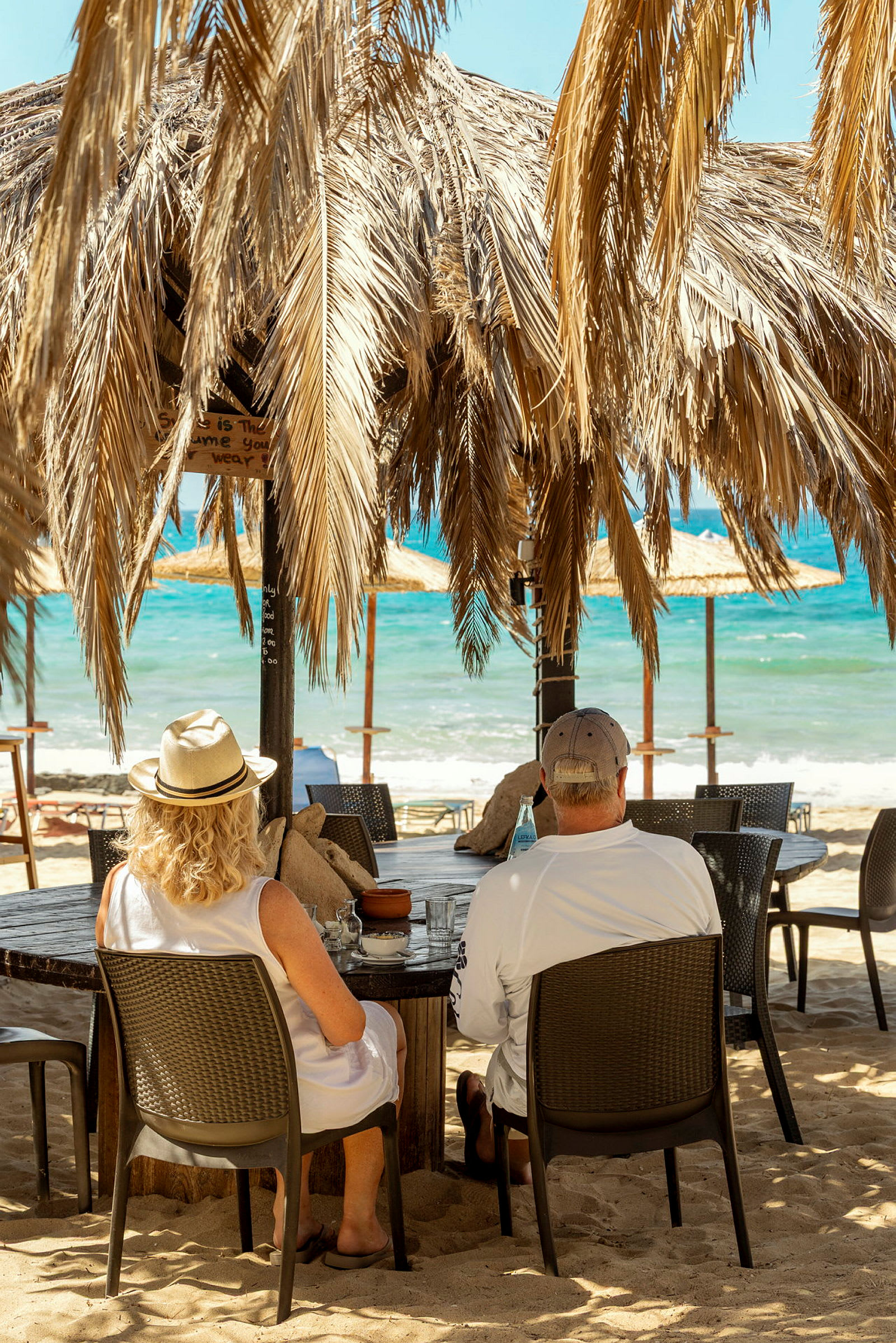 Två personer sitter vid ett bord under ett parasoll gjort av palmblad på en strand nära havet. (AI-genererad text)