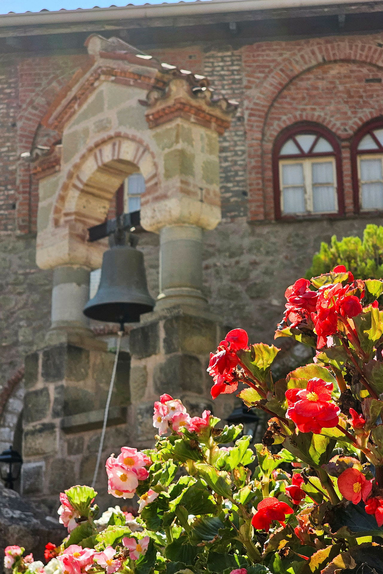 Bild på en kyrkobyggnad med en klocka i förgrunden och röda blommor i fokus. (AI-genererad text)