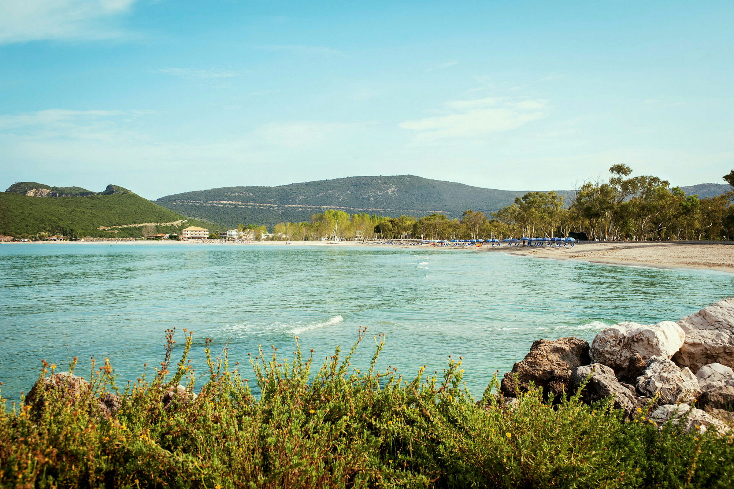 En strand med klart vatten, gröna kullar och träd i bakgrunden. (AI-genererad text)