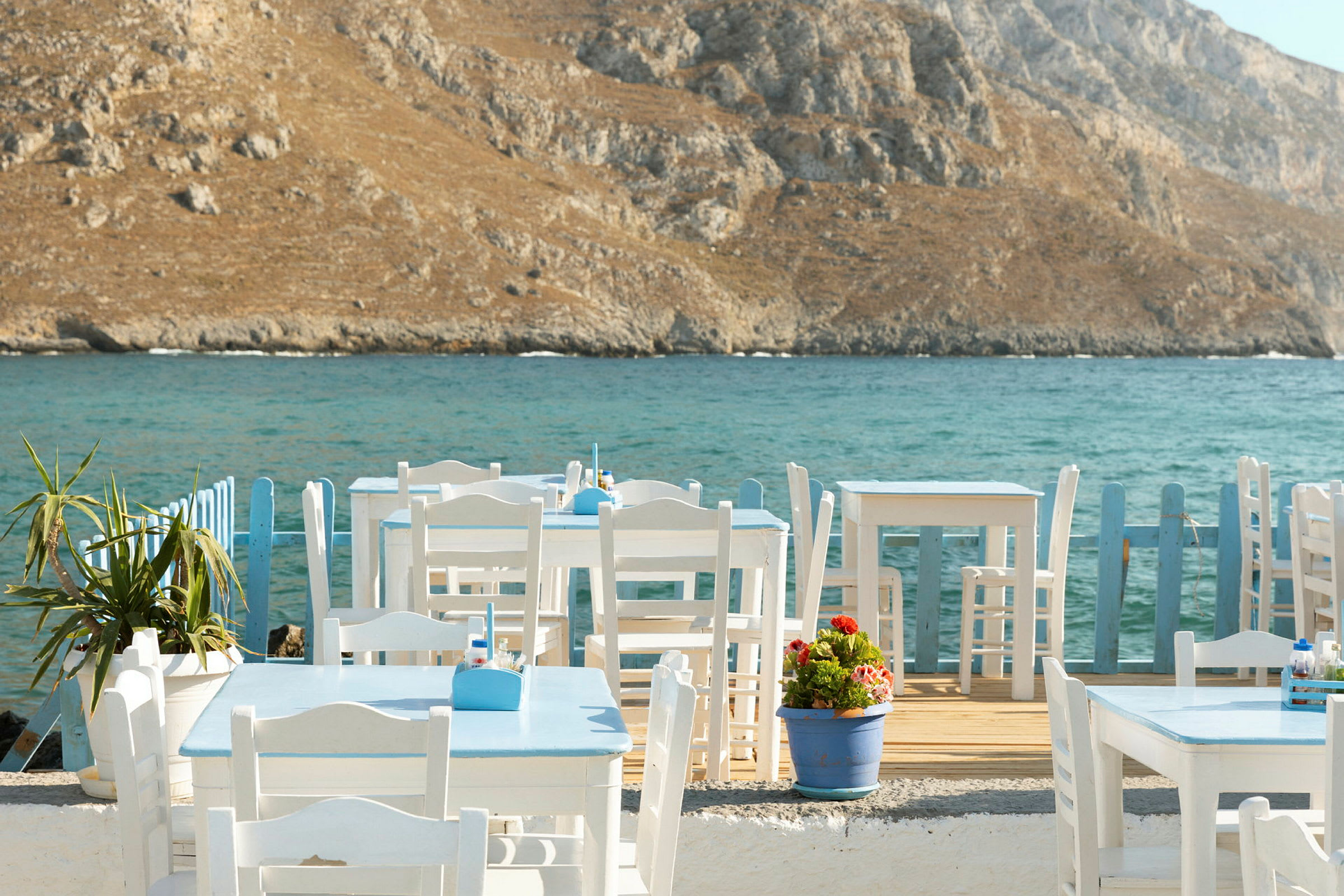 Vitmålade bord och stolar vid havet med klippig bakgrund. (AI-genererad text)