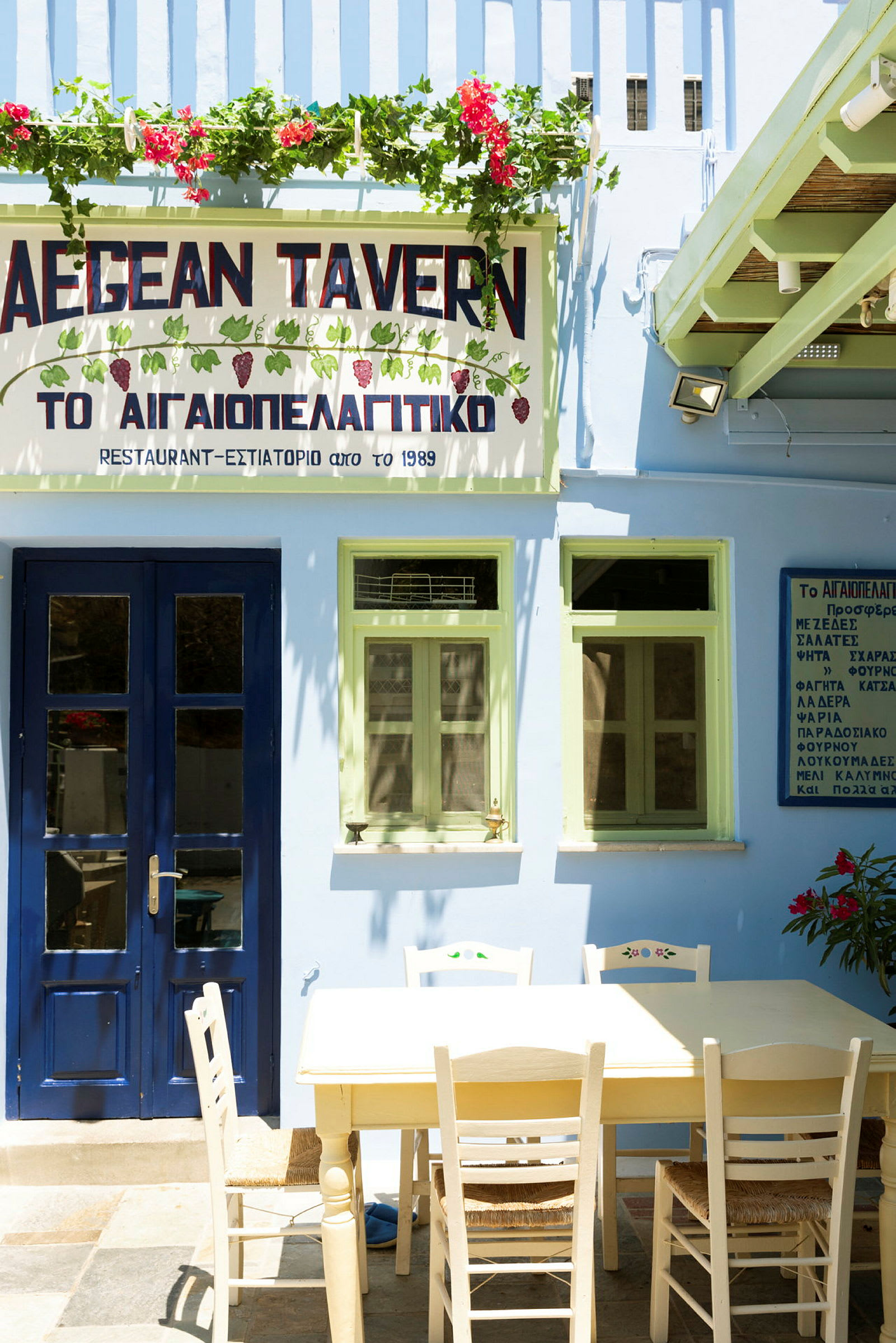 En byggnad med blå dörr och gröna fönster, en skylt med texten 'AEGEAN TAVERN' och ett bord med stolar framför. (AI-genererad text)