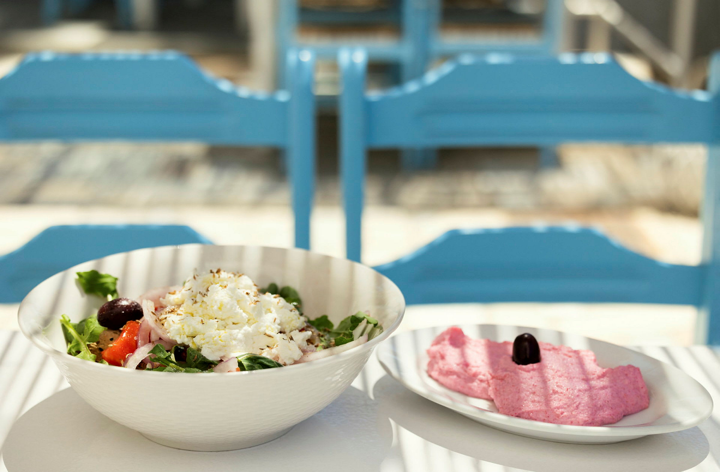 En skål med sallad och tallrik med rosa röra samt en oliv på ett bord med blå stolar i bakgrunden. (AI-genererad text)