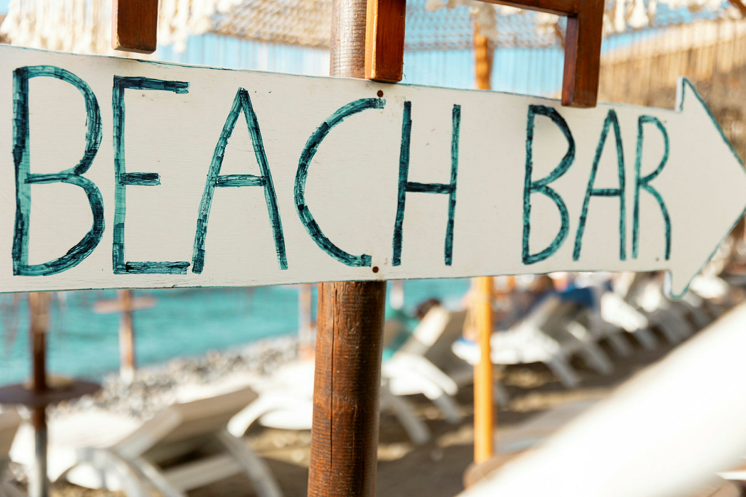 Skylt med texten 'BEACH BAR' och en pil, i bakgrunden stranden med solstolar och havet. (AI-genererad text)