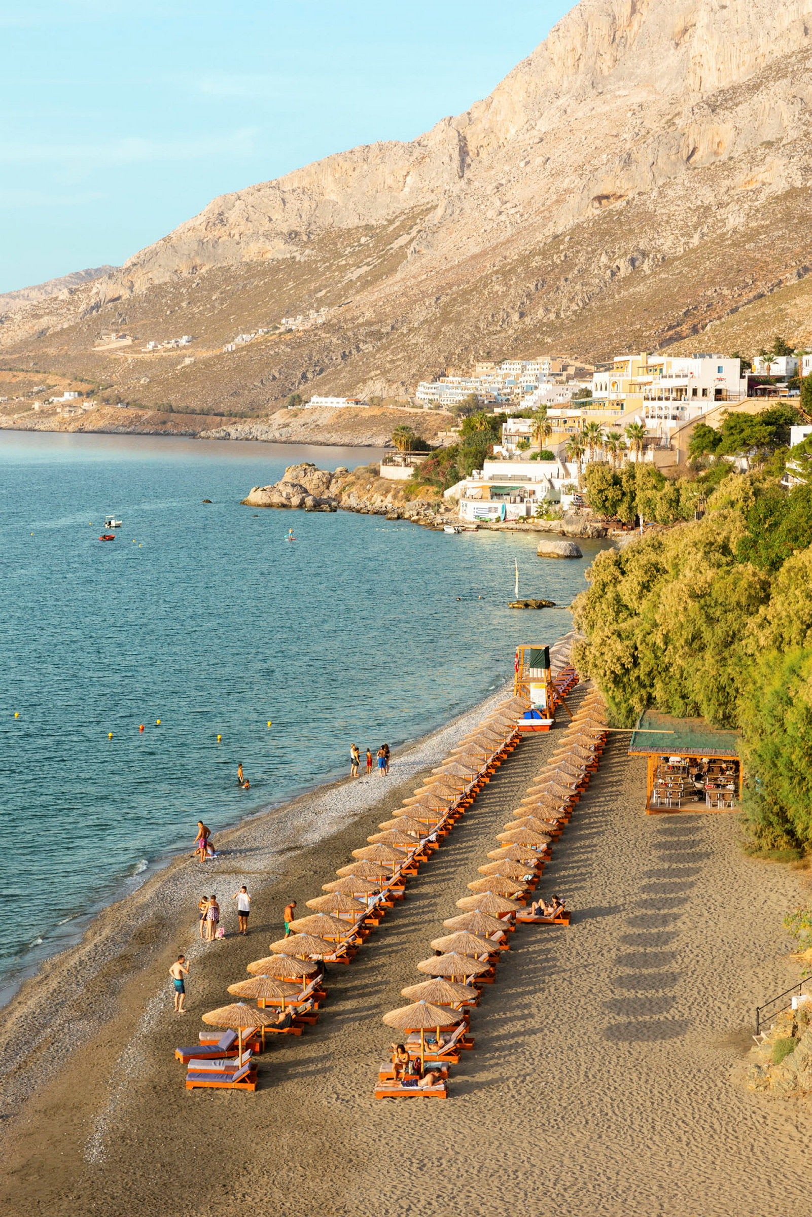 Strand med solstolar och parasoller längs vattenkanten, omgivna av berg och byggnader. (AI-genererad text)