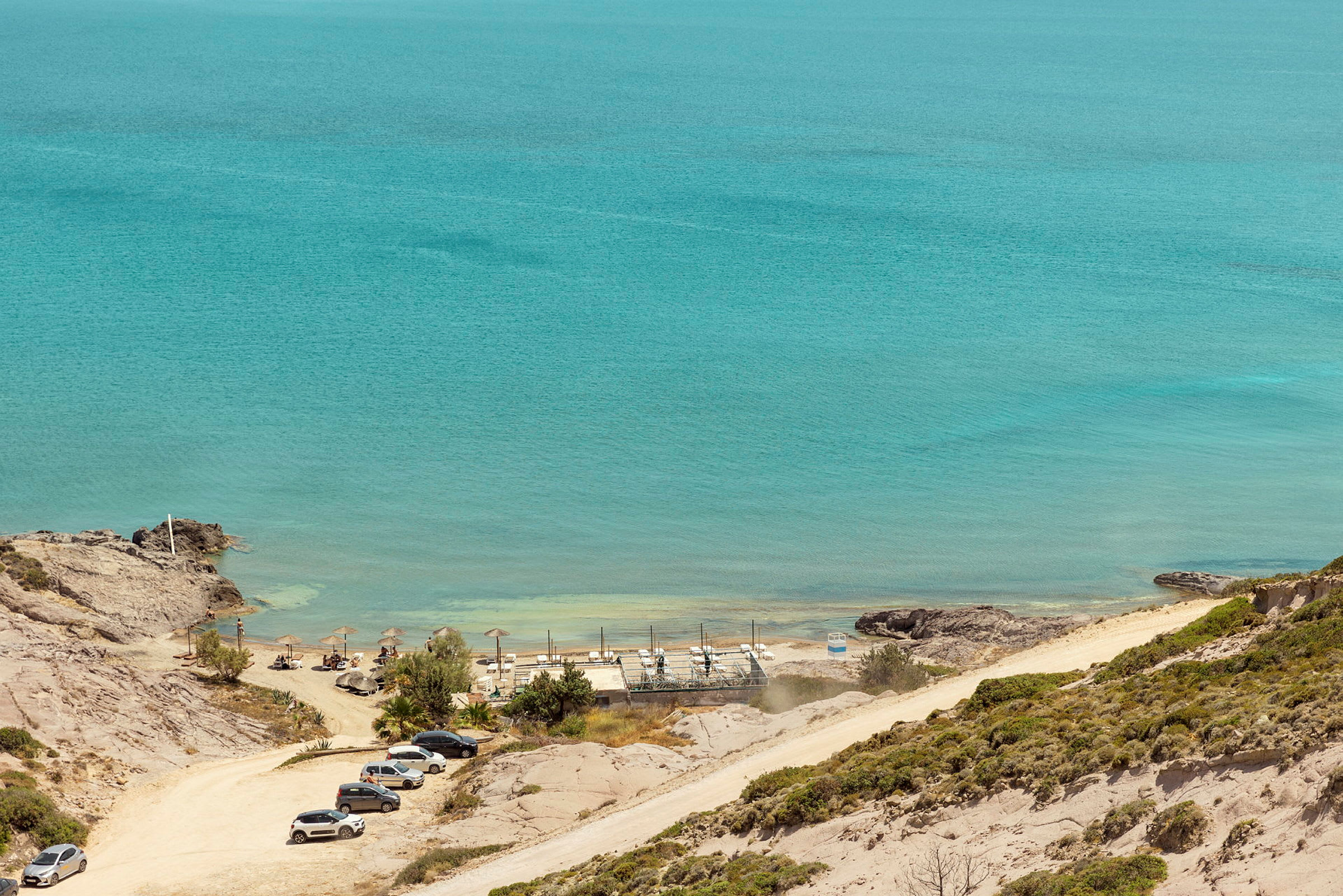 Strand med parasoller, solstolar och parkerade bilar, havet i bakgrunden. (AI-genererad text)