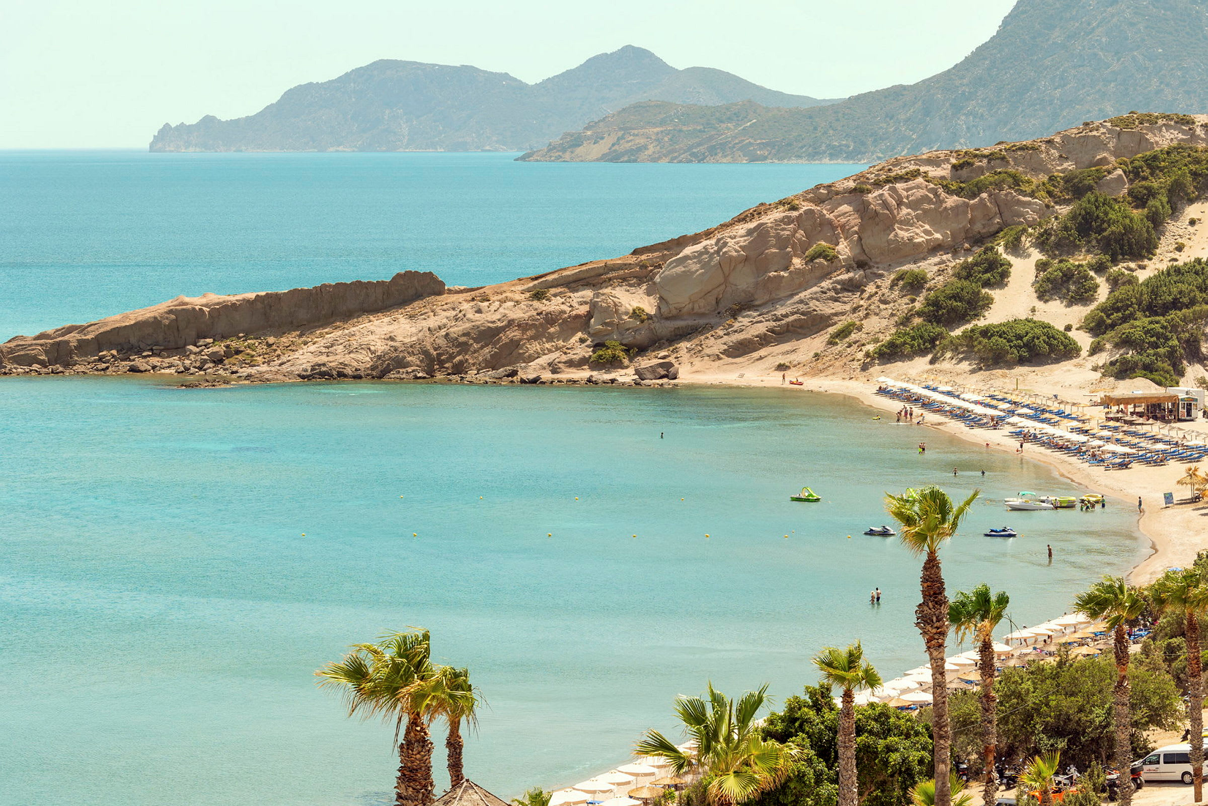 Strand med parasoller och solstolar vid en bukt med klart vatten, omgiven av klippor och vegetation. (AI-genererad text)