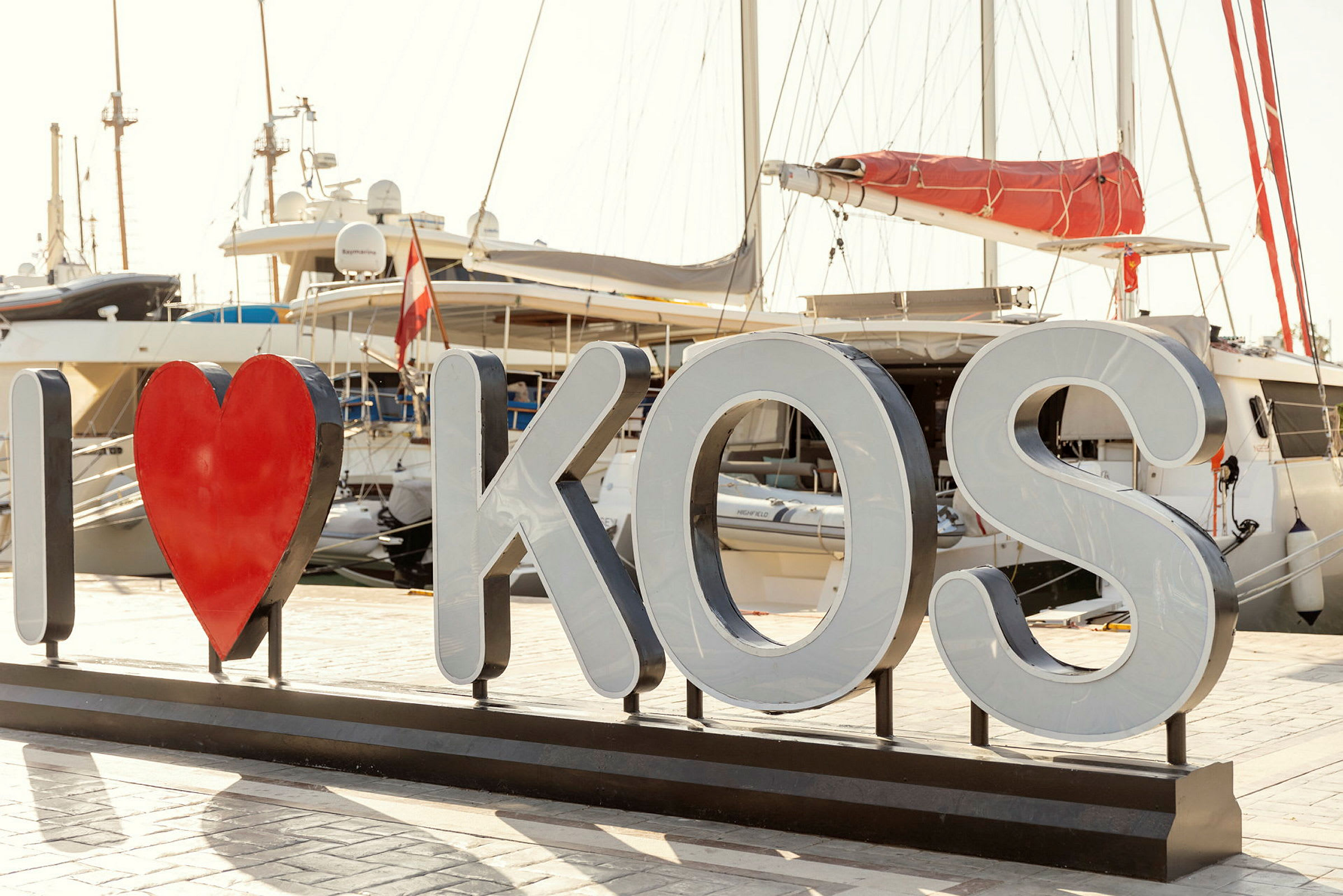 Skylt med texten 'I ♥ KOS' framför båtar i en hamn. (AI-genererad text)