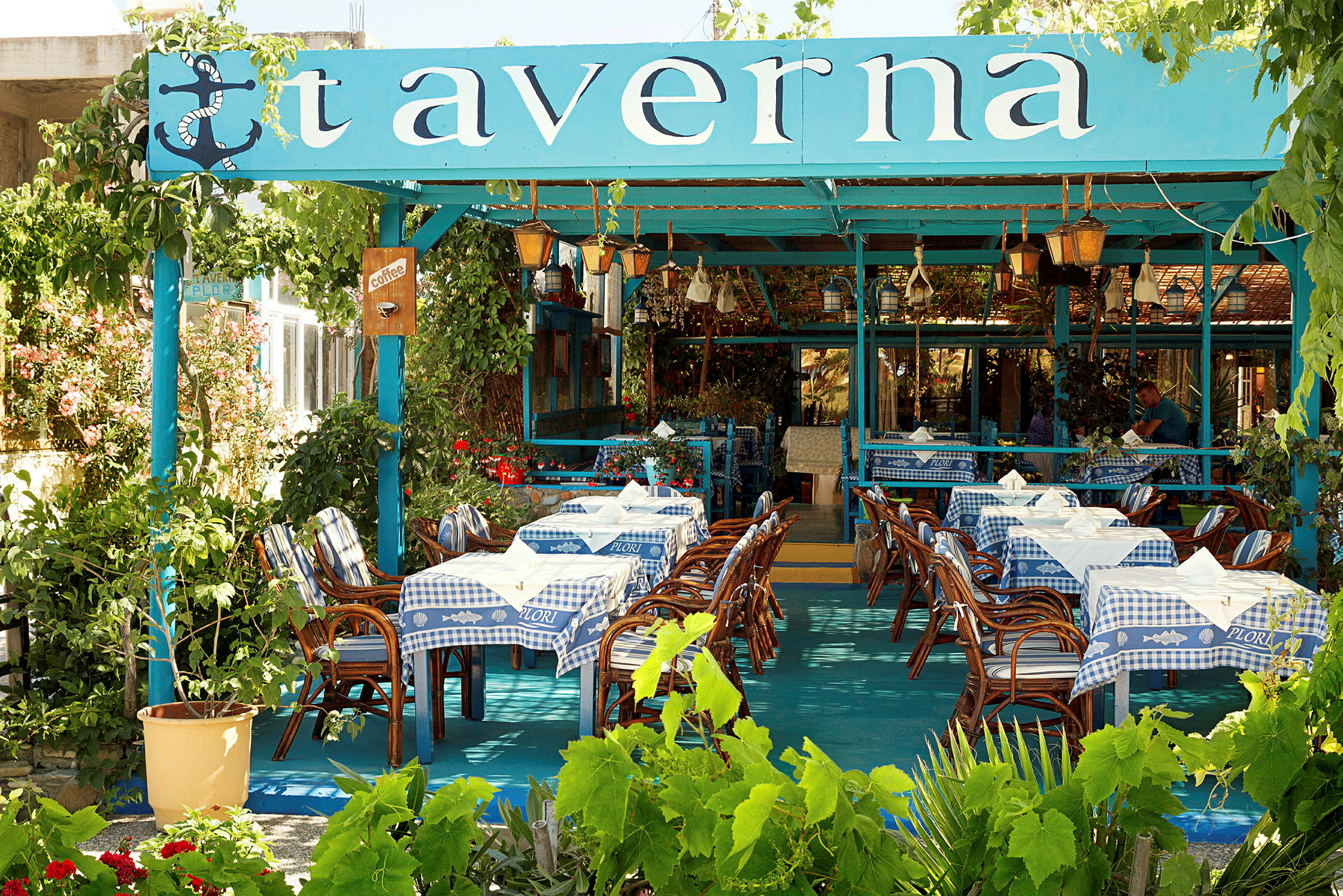 En traditionell taverna med blå och vita dukar, bord och stolar i trä, samt grönska omkring. (AI-genererad text)