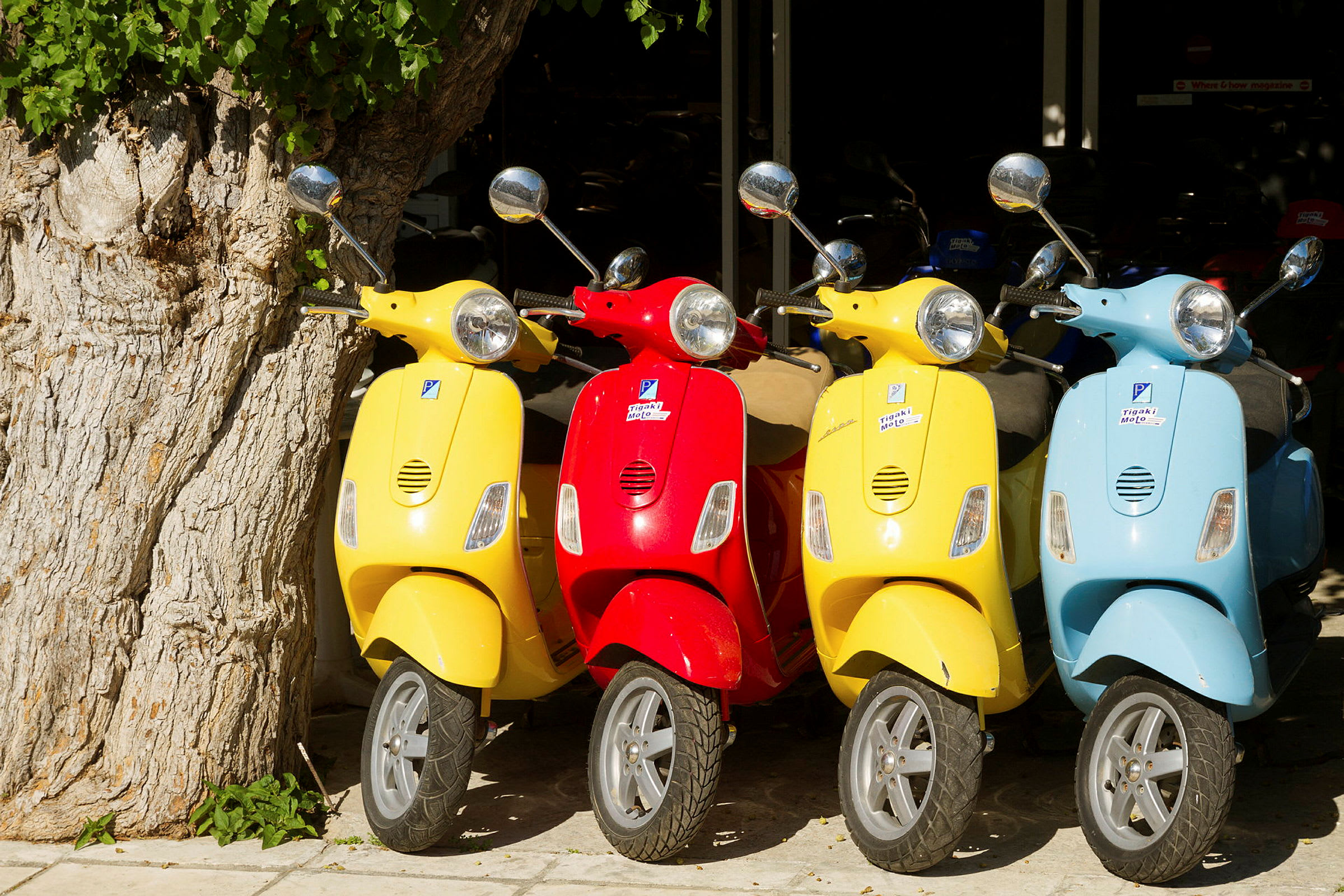 Flera färgglada scooters, röda, gula och blå, parkerade bredvid ett träd. (AI-genererad text)
