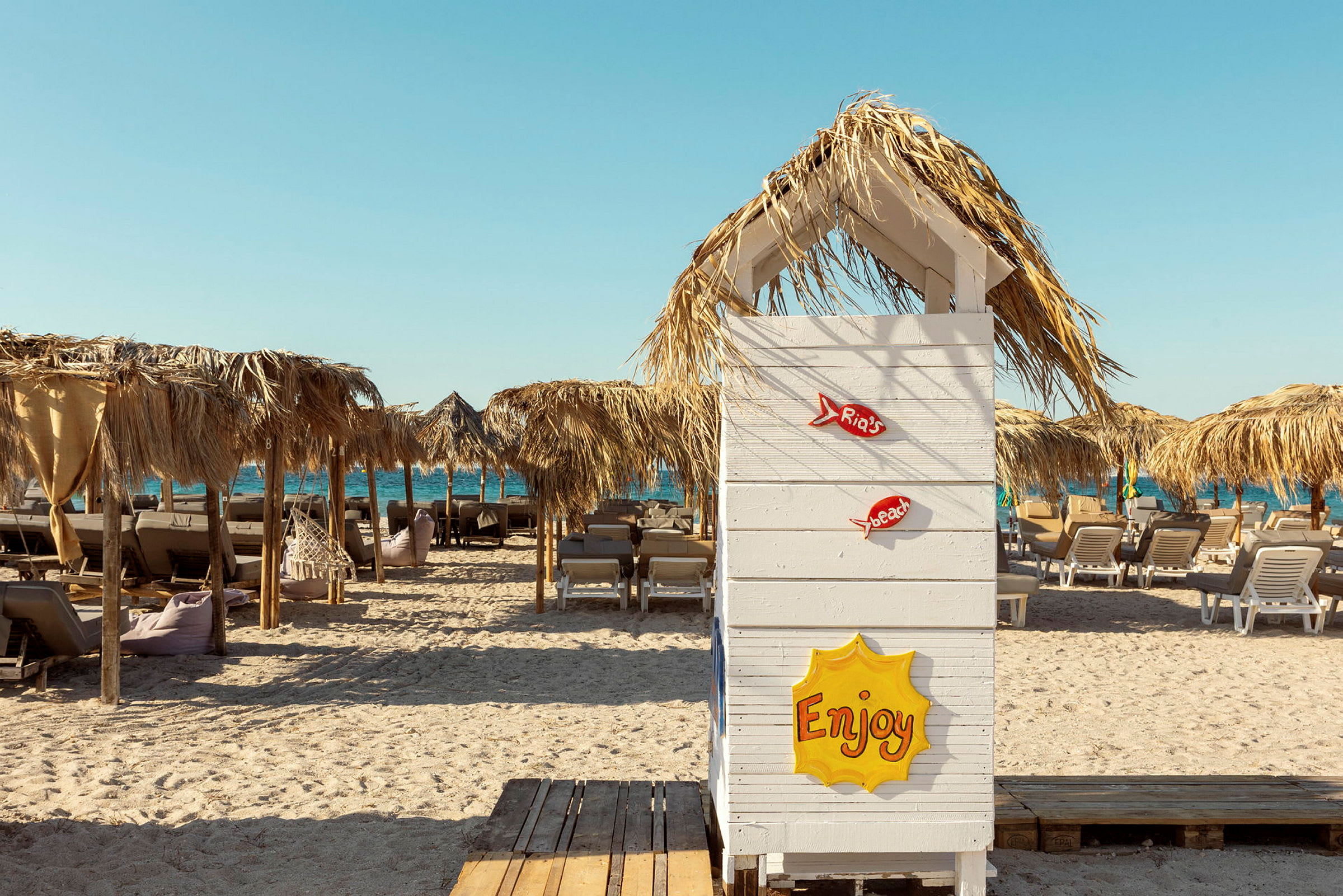 En strand med solstolar och parasoller av halm samt en vit träskärm närmast i bild med texten ”Enjoy”. (AI-genererad text)