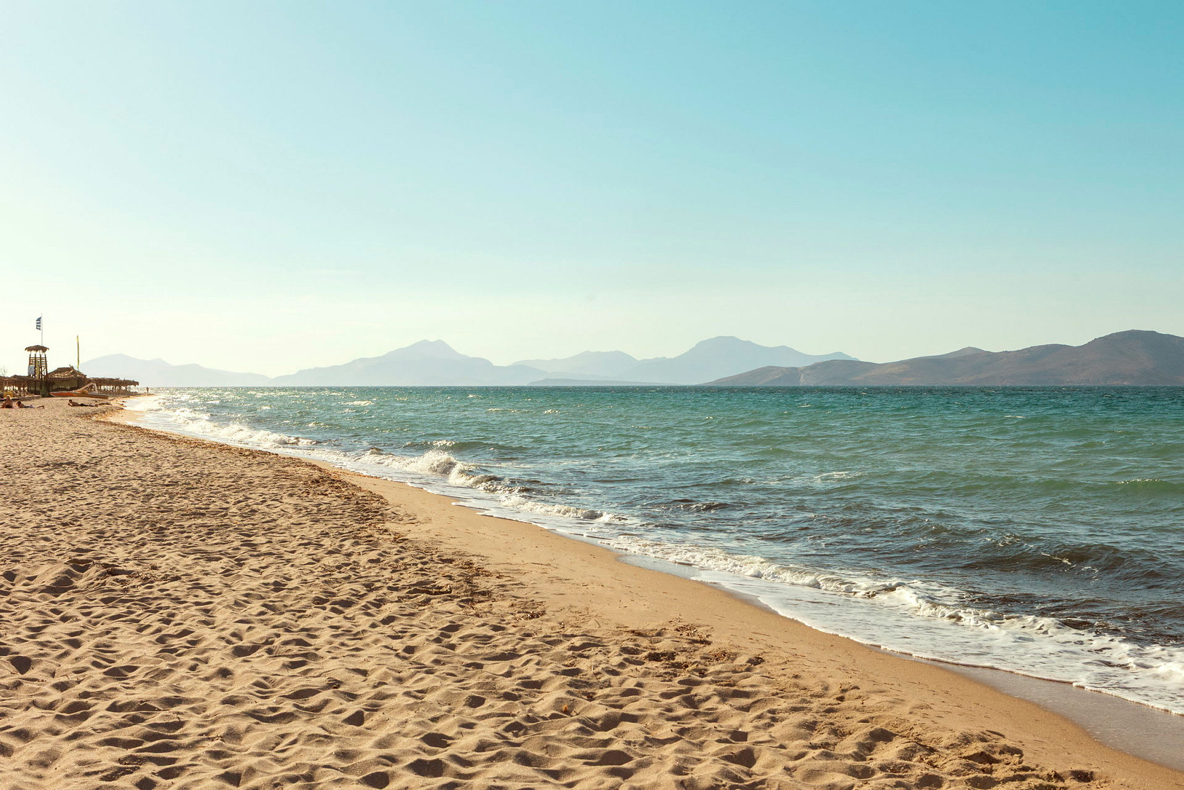 En solbelyst sandstrand med vågigt hav i bakgrunden. (AI-genererad text)
