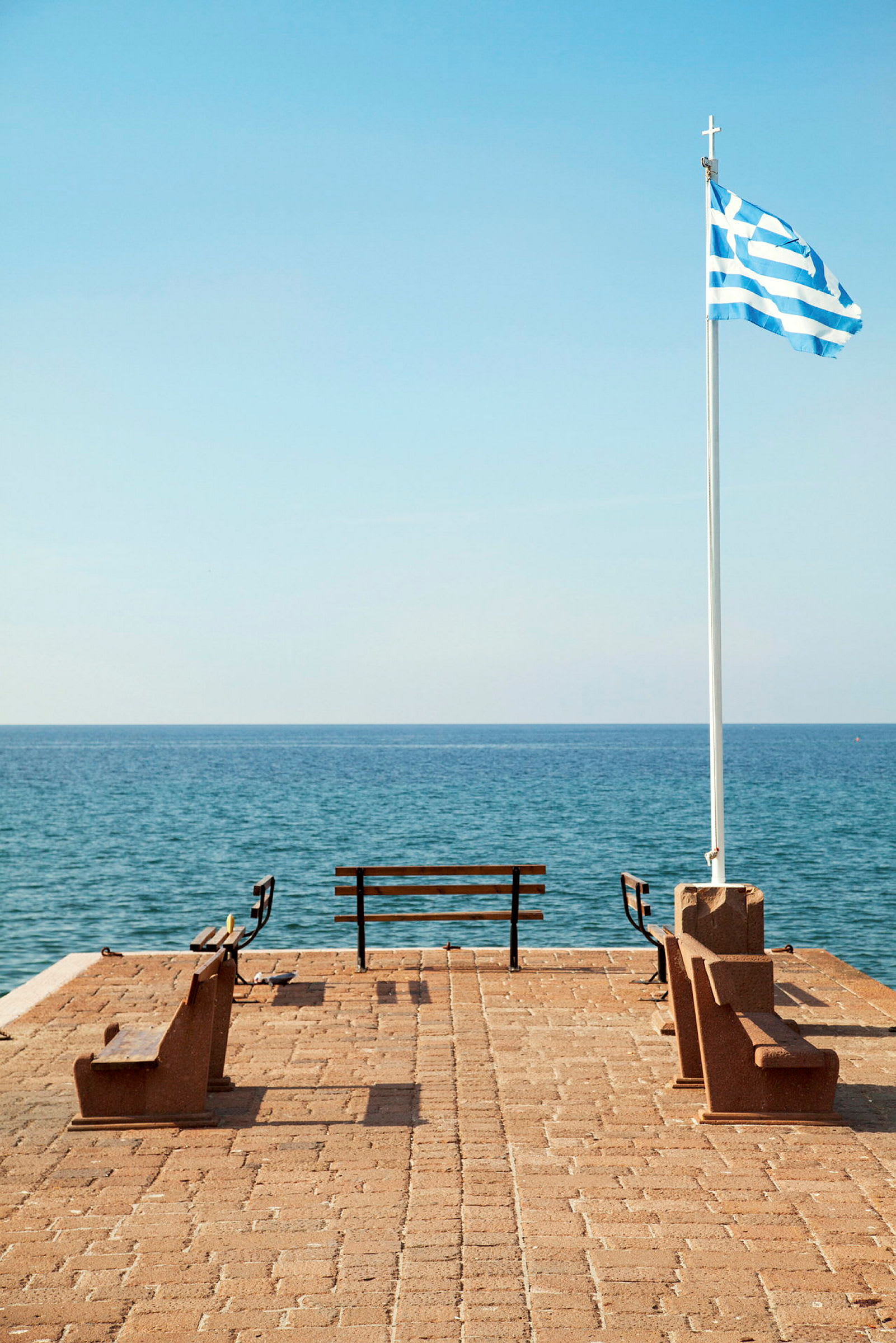 Brygga med bänkar och grekisk flagga vid havet. (AI-genererad text)
