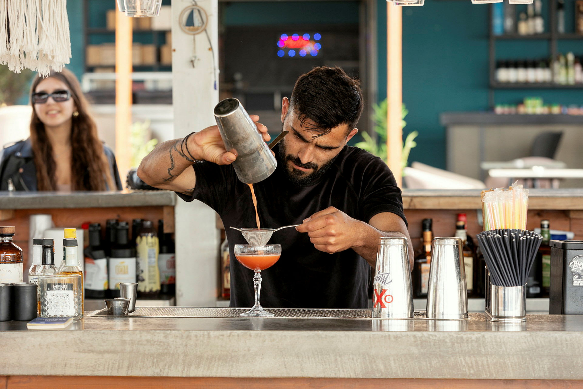 En bartender skakar och silar en cocktail bredvid flaskor, stålshakers och sugrör på en utomhusbar. (AI-genererad text)
