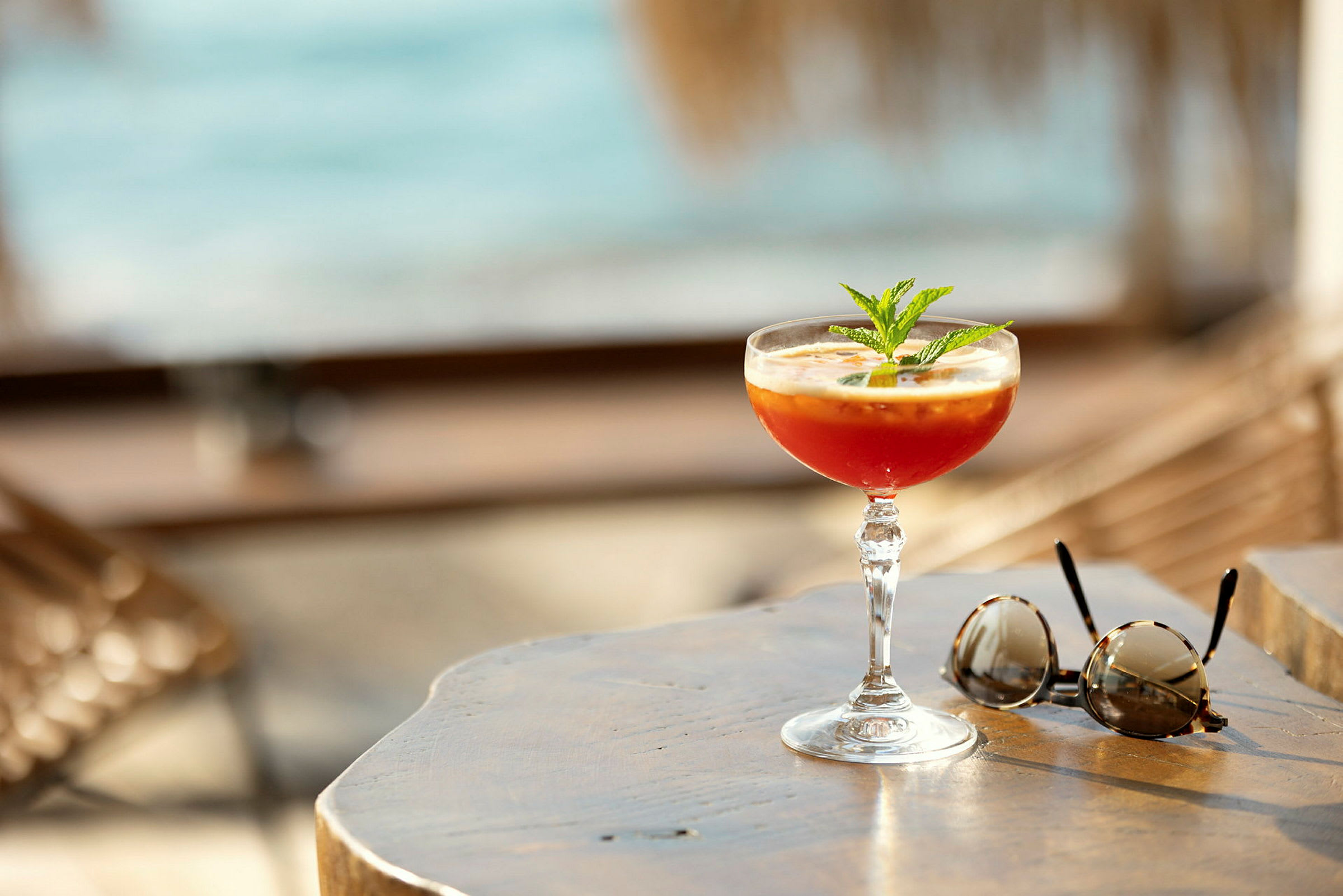 En cocktail och ett par solglasögon på ett träbord vid stranden. (AI-genererad text)