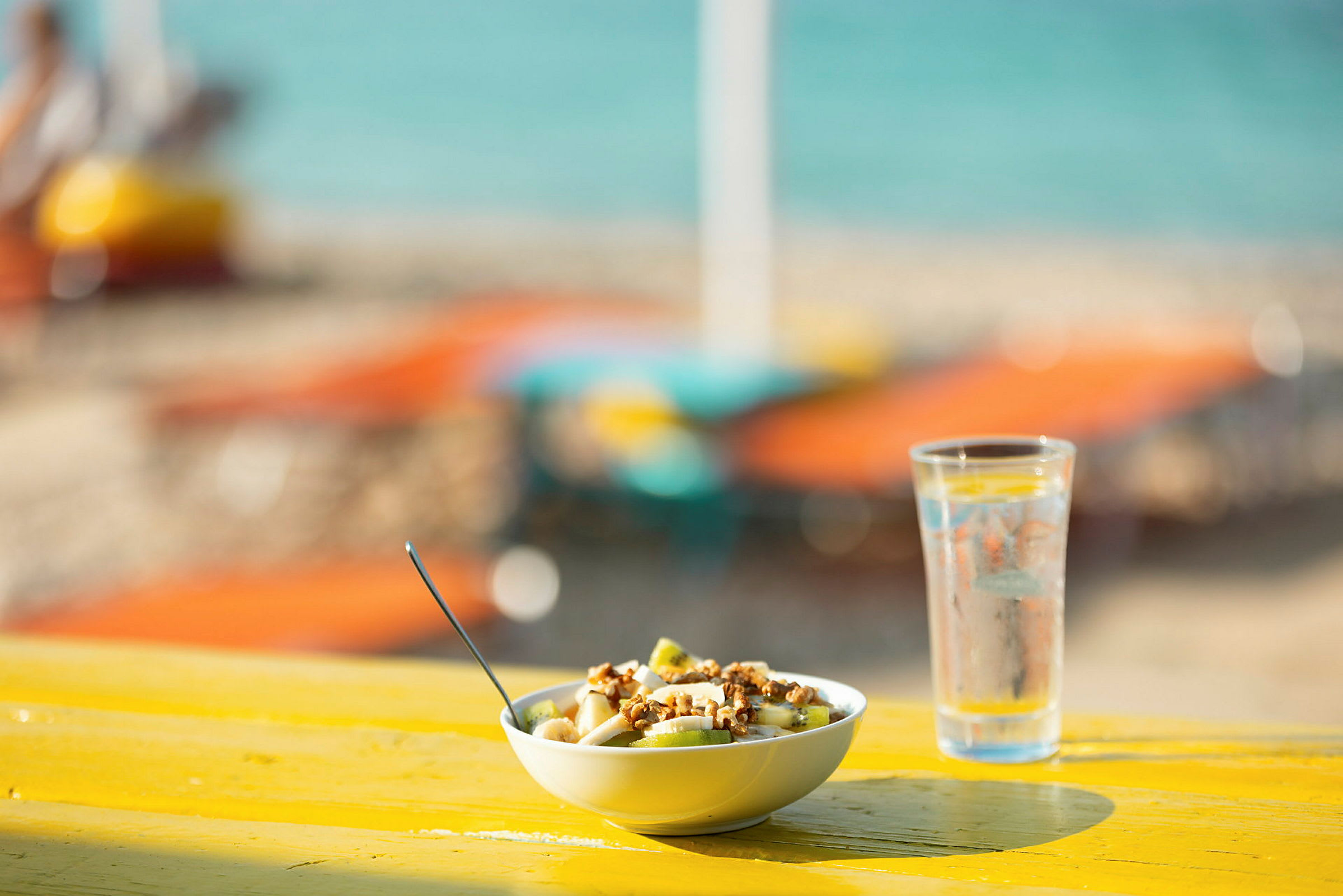 En skål med frukt och nötter bredvid ett glas vatten på ett gult bord vid stranden. (AI-genererad text)