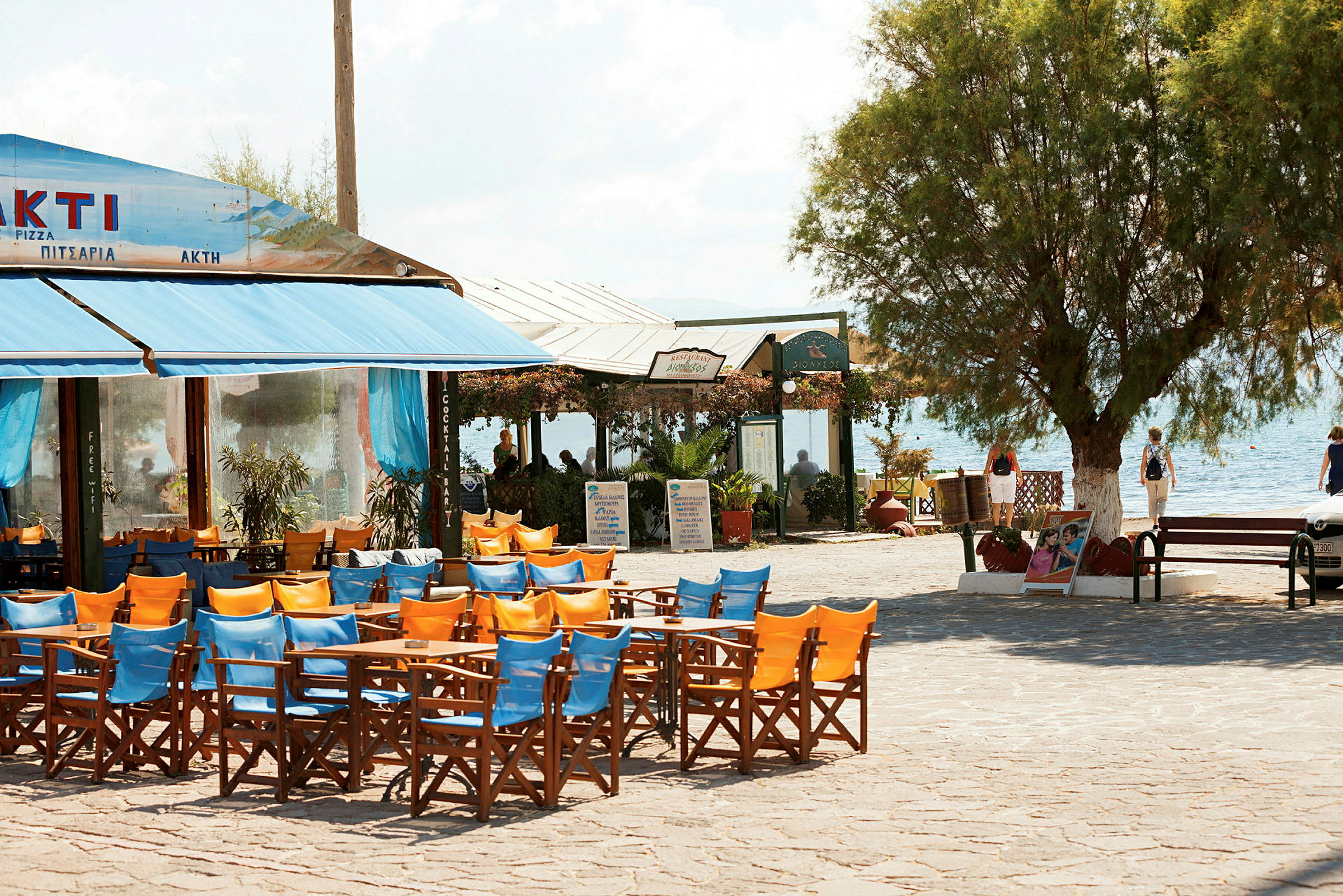 En utomhusrestaurang med färgglada stolar och bord med havet i bakgrunden. (AI-genererad text)