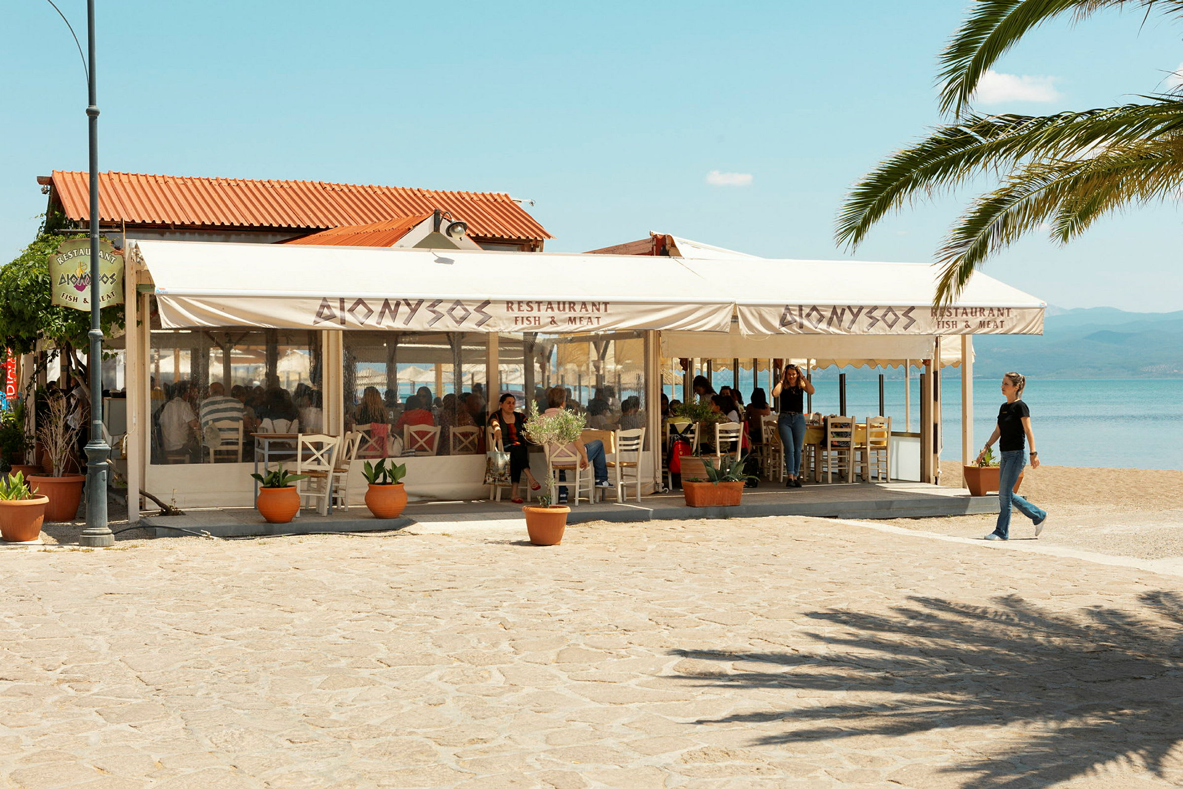 En restaurang vid stranden med skyltar som anger att den serverar fisk och kött. Uteserveringen är täckt med parasoller. (AI-genererad text)
