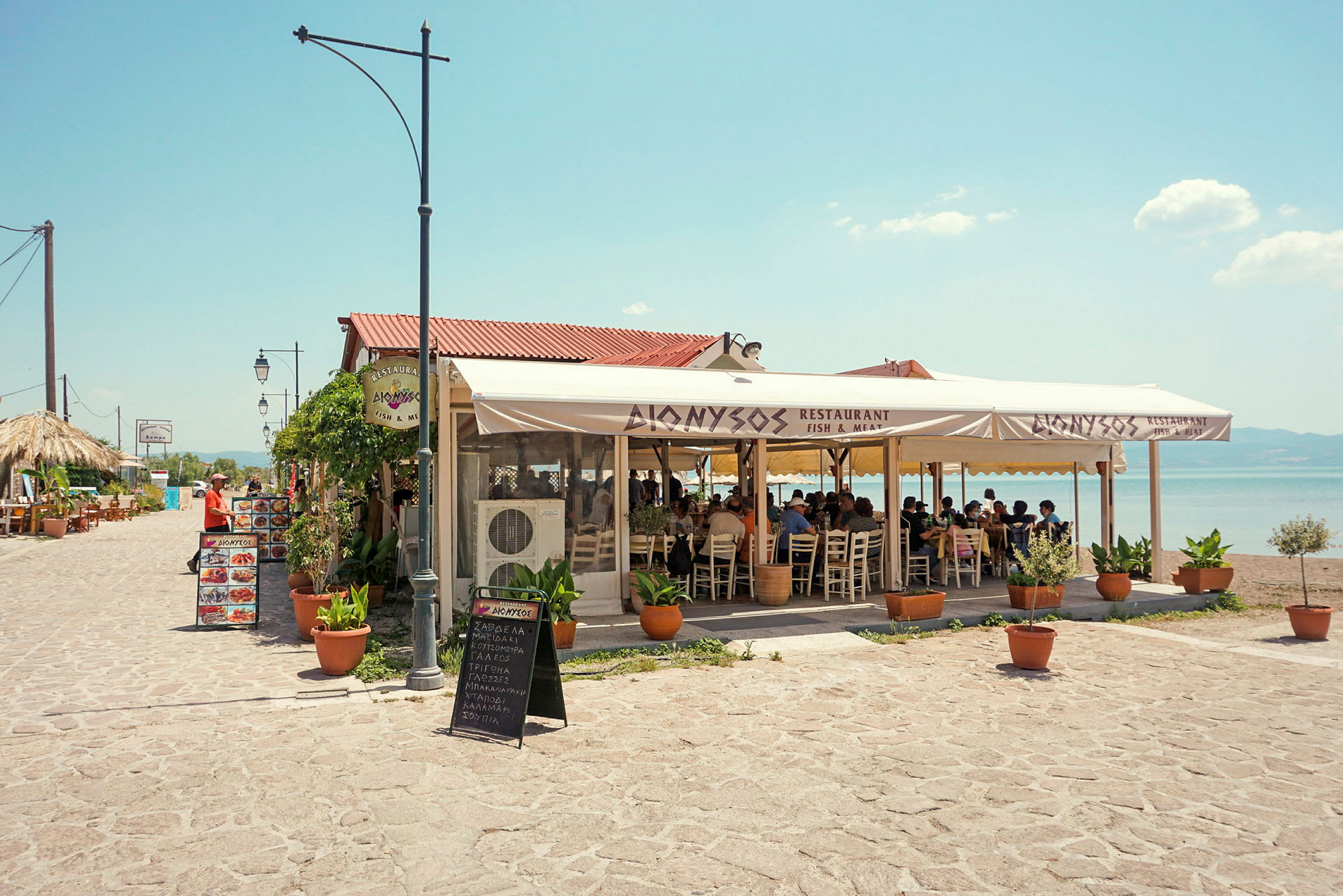 En utomhusrestaurang med sittplatser under ett stort tält. I bakgrunden syns havet och en klar himmel. (AI-genererad text)