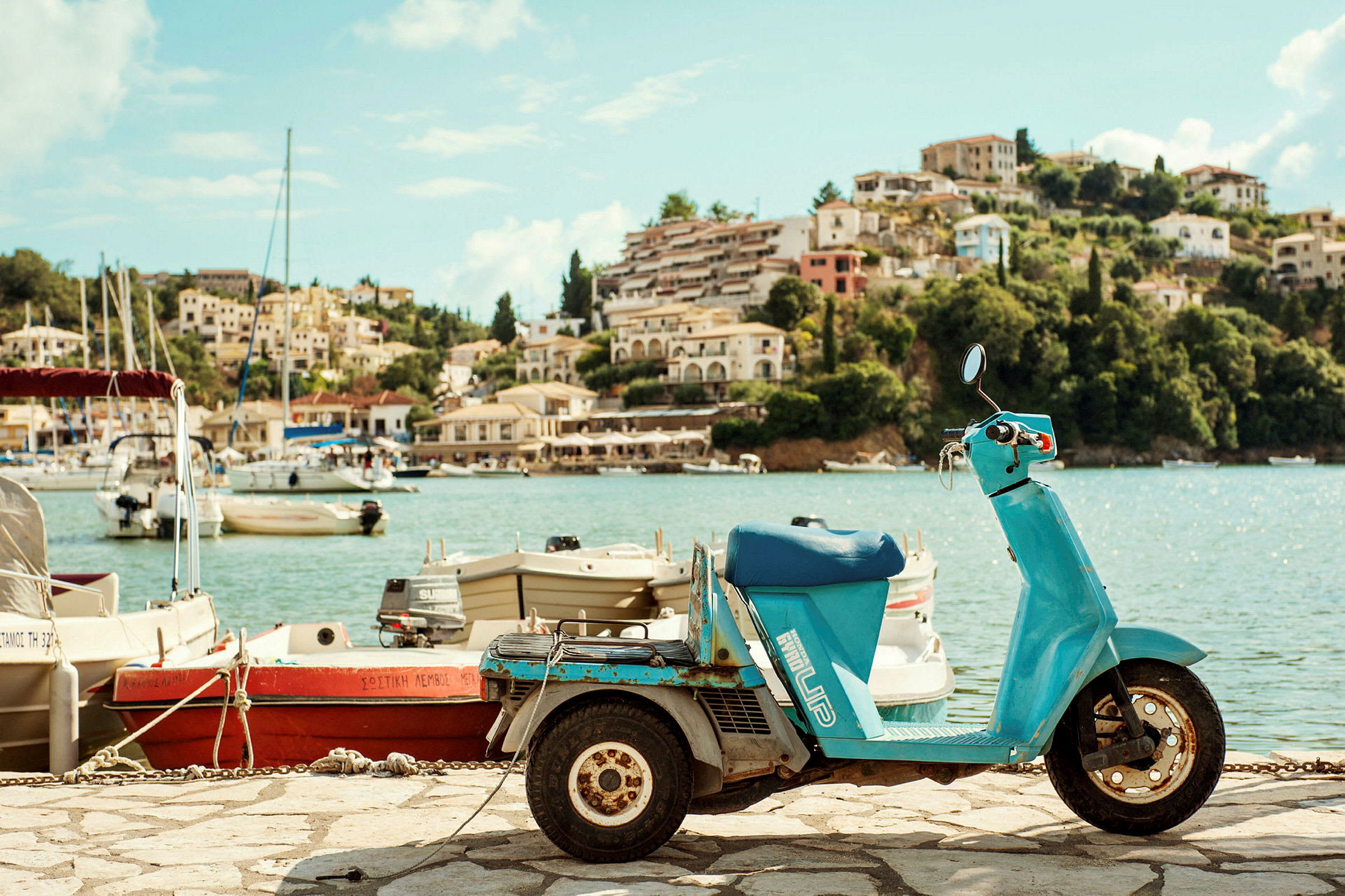 En blå scooter står parkerad vid en hamn med båtar och en bergssluttning i bakgrunden. (AI-genererad text)