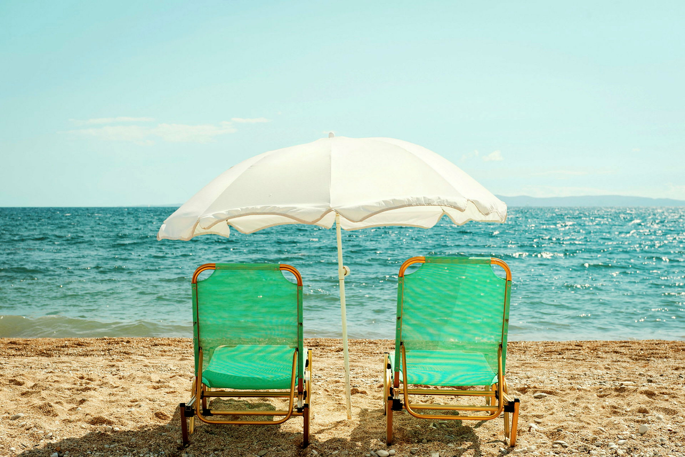 Två solsängar med gröna sittdynor och ett vitt parasoll står på stranden framför havet. (AI-genererad text)