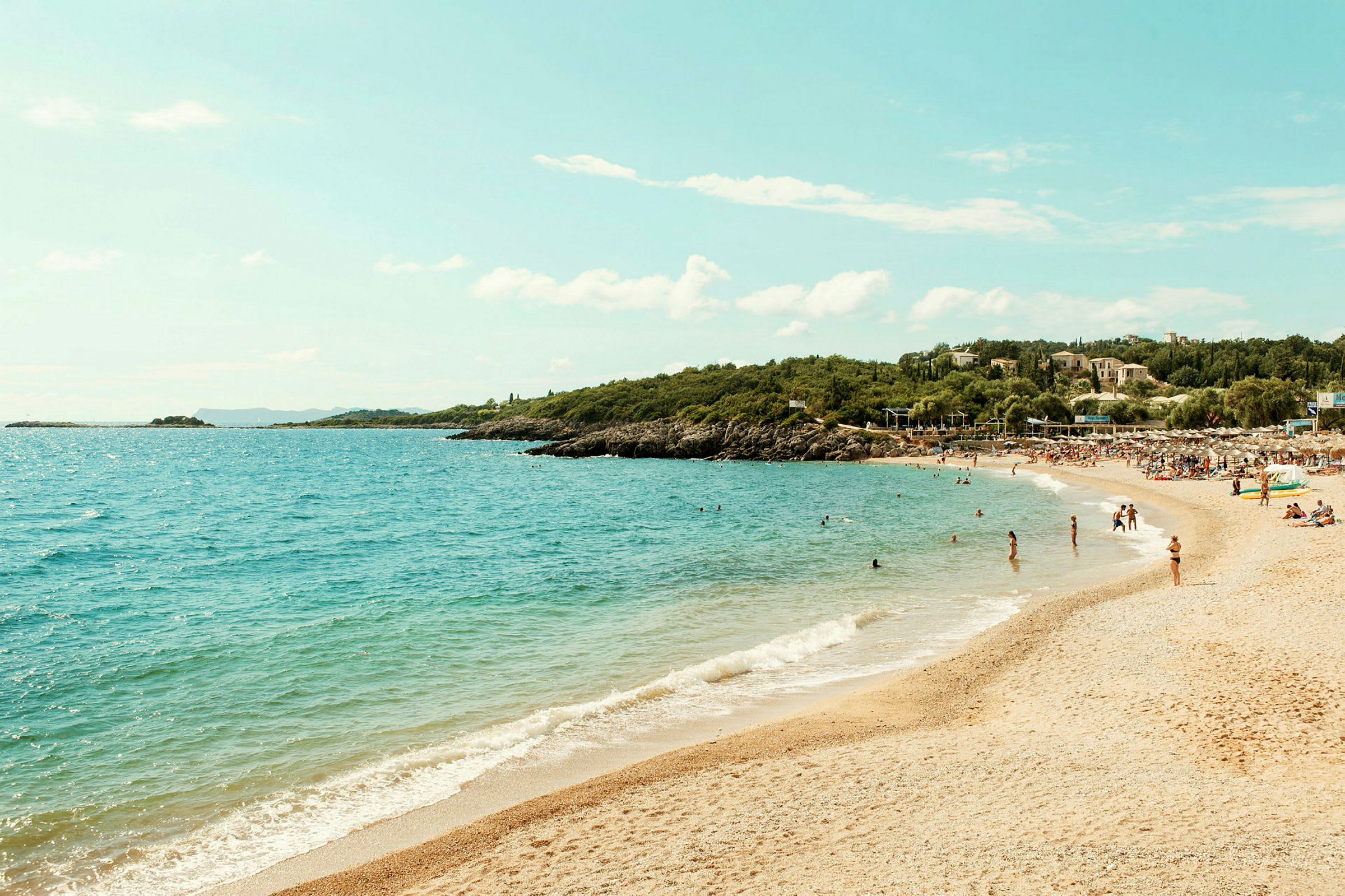 Strand med sand och klart blått vatten, omgiven av grönska och människor som badar. (AI-genererad text)
