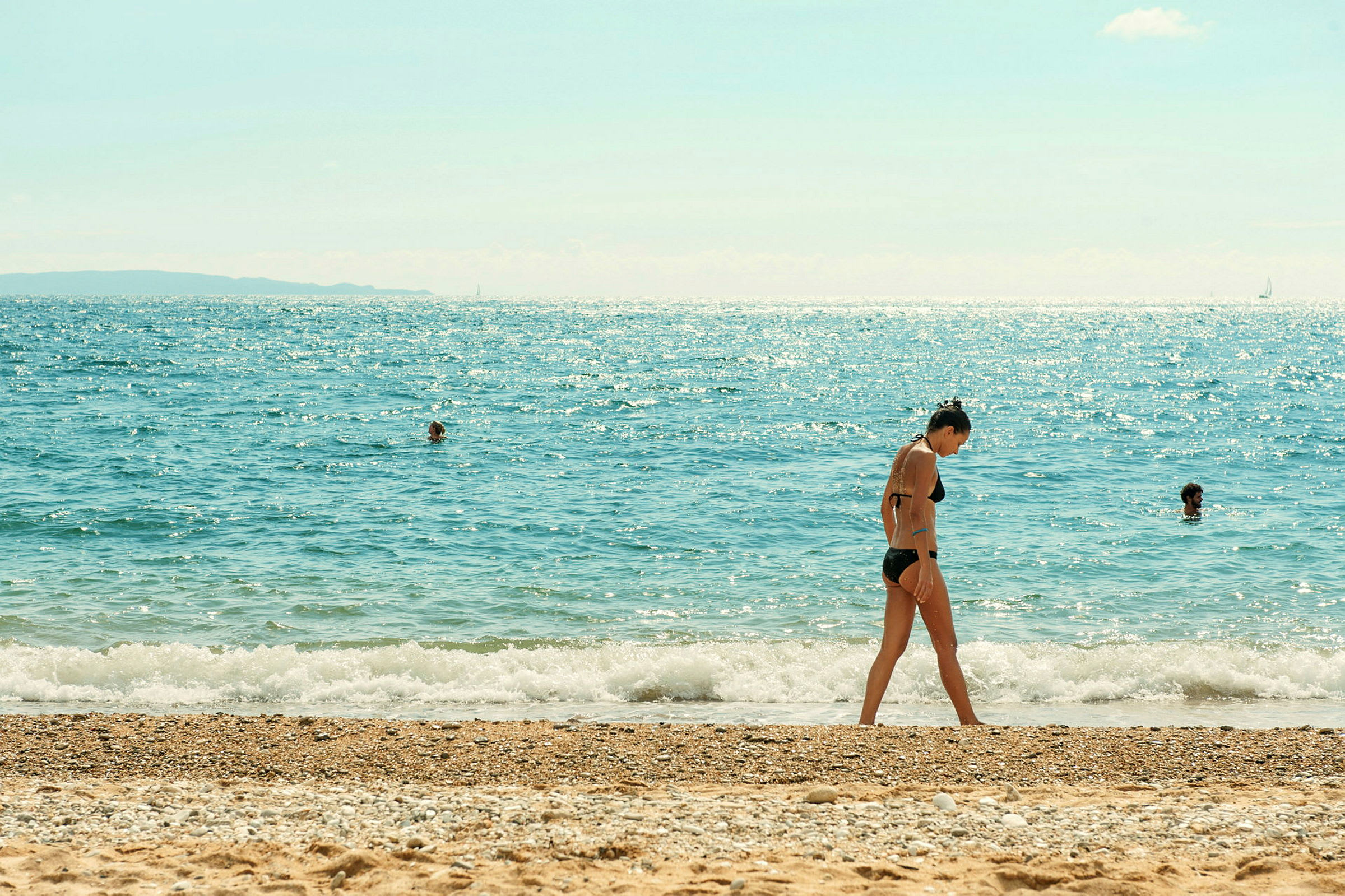 En strand med sand och sten, och havet i bakgrunden med personer som badar. (AI-genererad text)