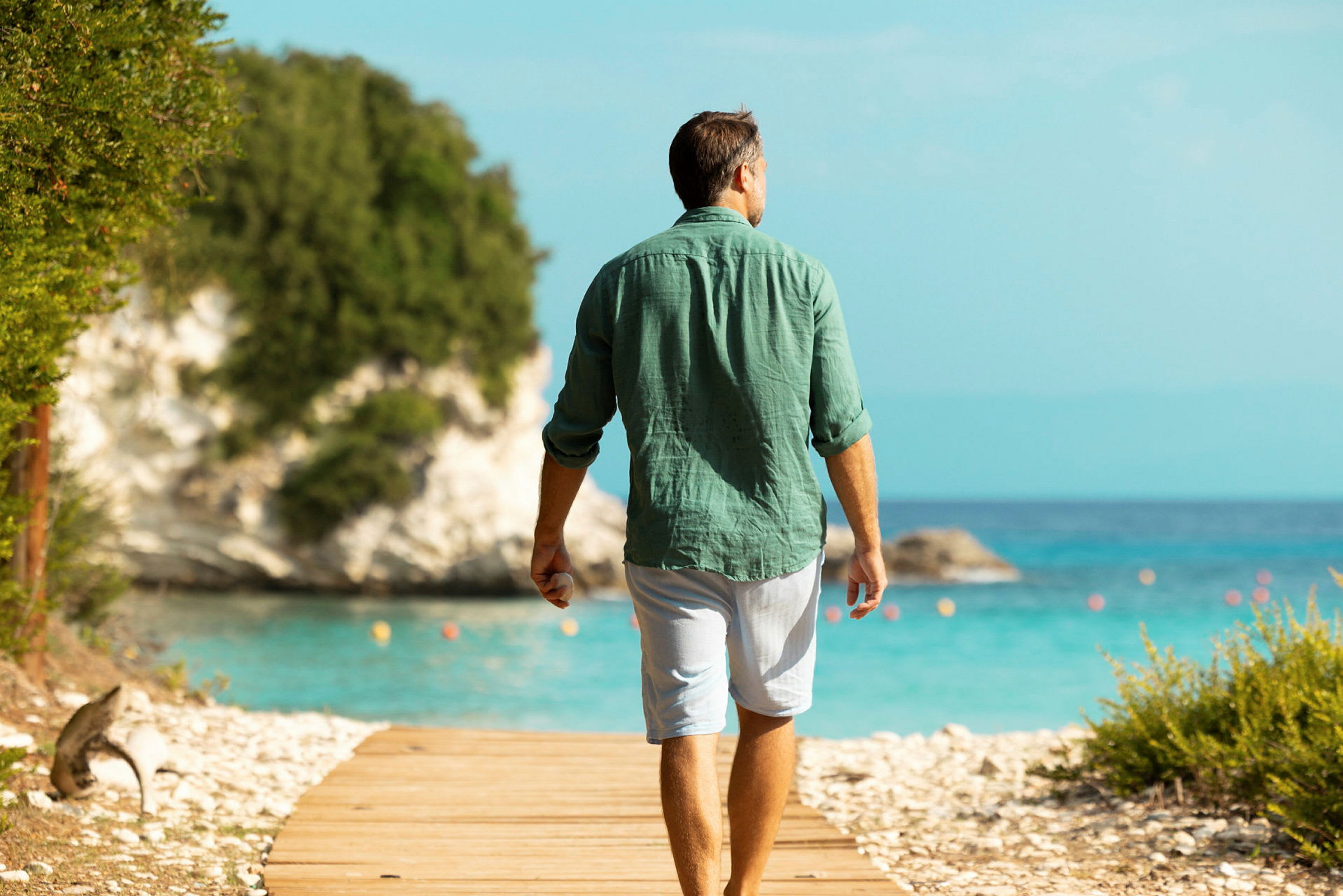 En person går på en trästig bredvid havet med grönskande klippor i bakgrunden. (AI-genererad text)