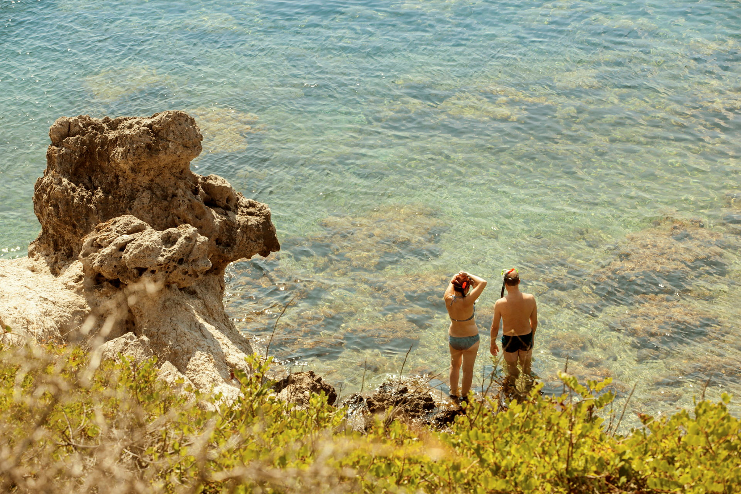 Två personer står i klart vatten vid en klippa, utrustade med snorkelutrustning. (AI-genererad text)