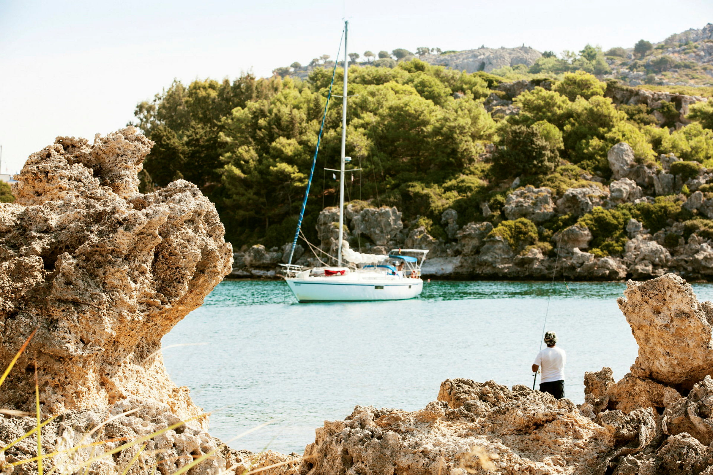 En segelbåt ligger förtöjd vid en rofylld vik omgiven av klippor och skog med en person som fiskar från stranden. (AI-genererad text)