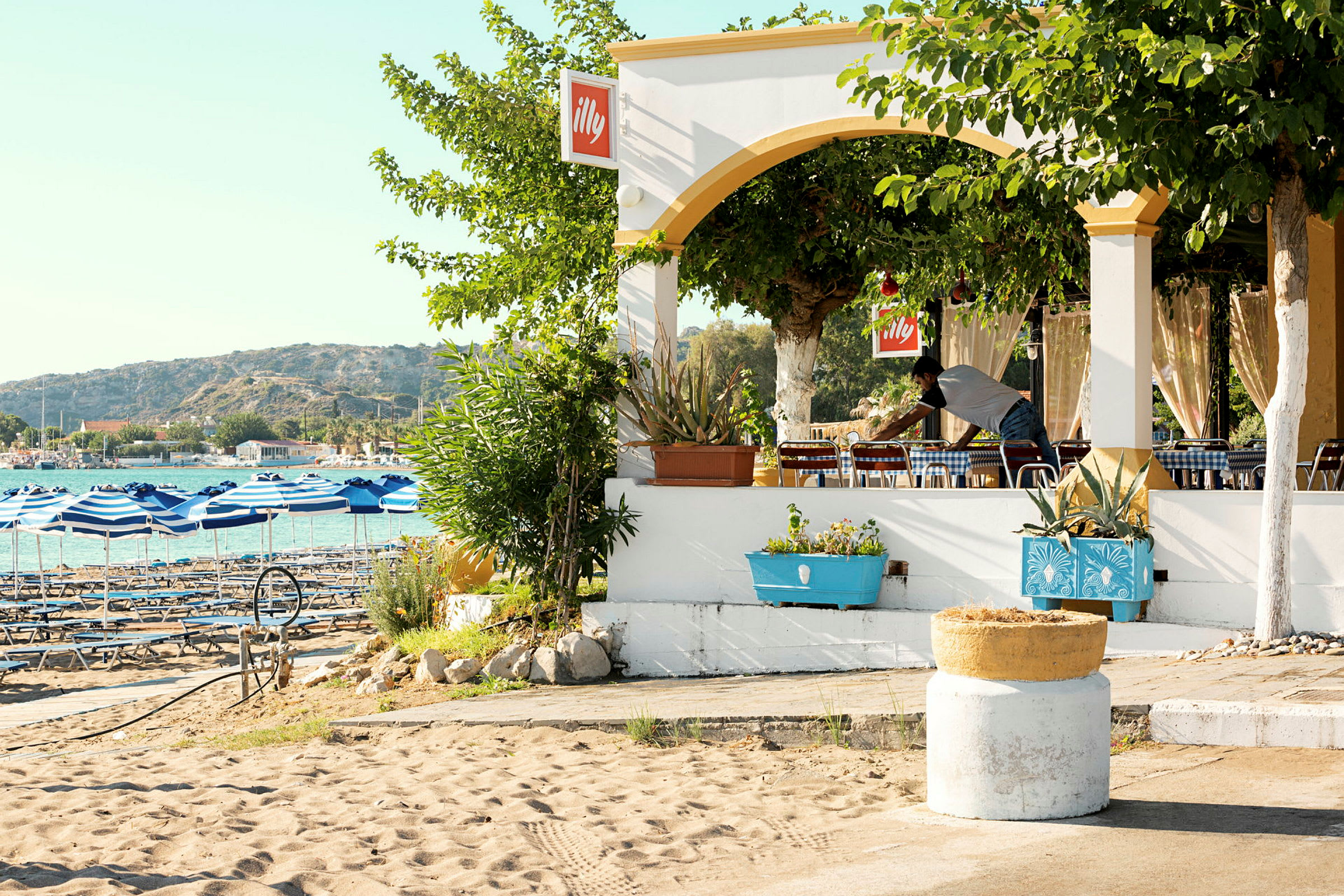 En strandnära restaurang med blåvitrandiga parasoller vid havet och omgivande grönska. (AI-genererad text)
