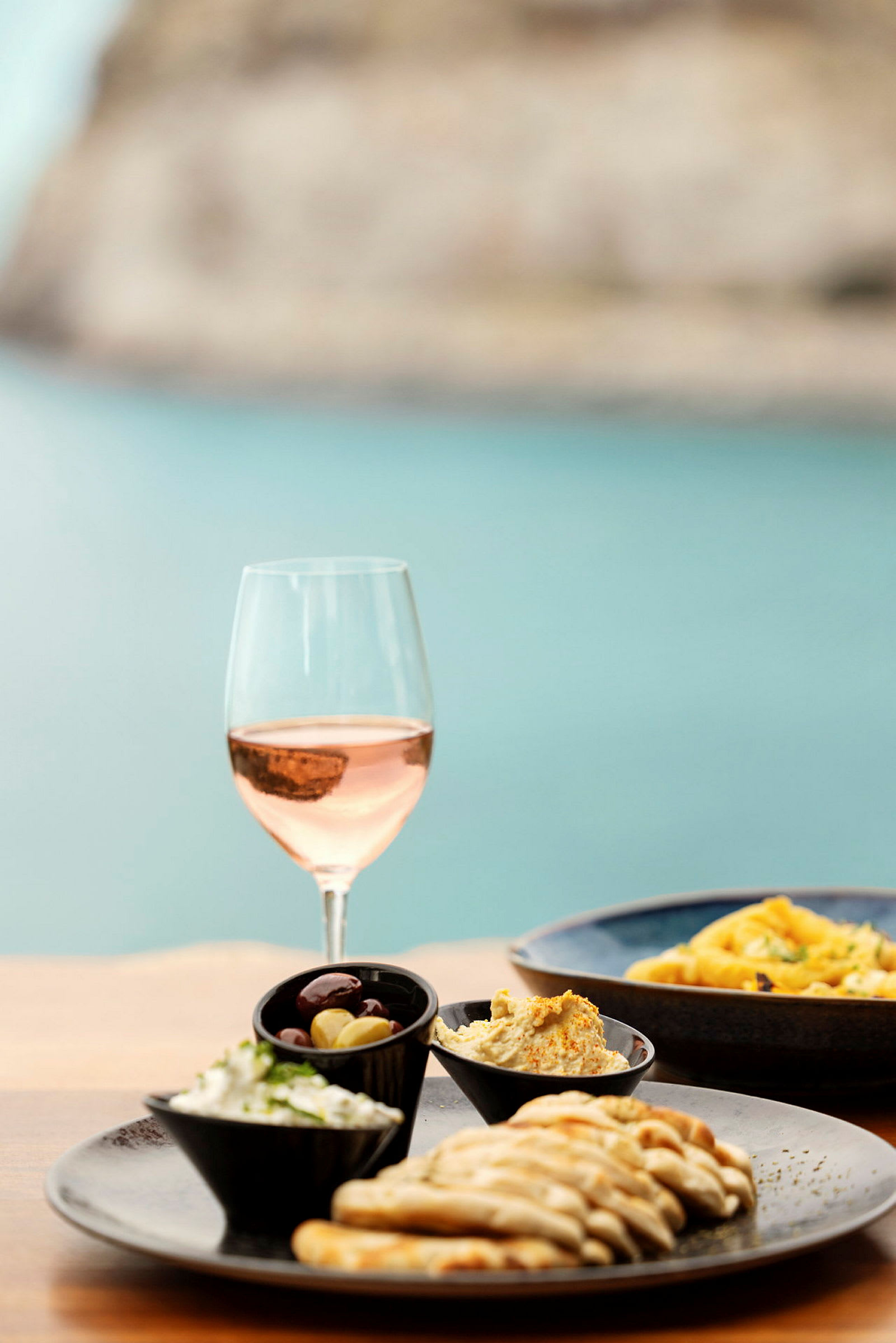 Ett glas rosévin bredvid en tallrik med brödskivor, olivskål, och krämiga röror. (AI-genererad text)
