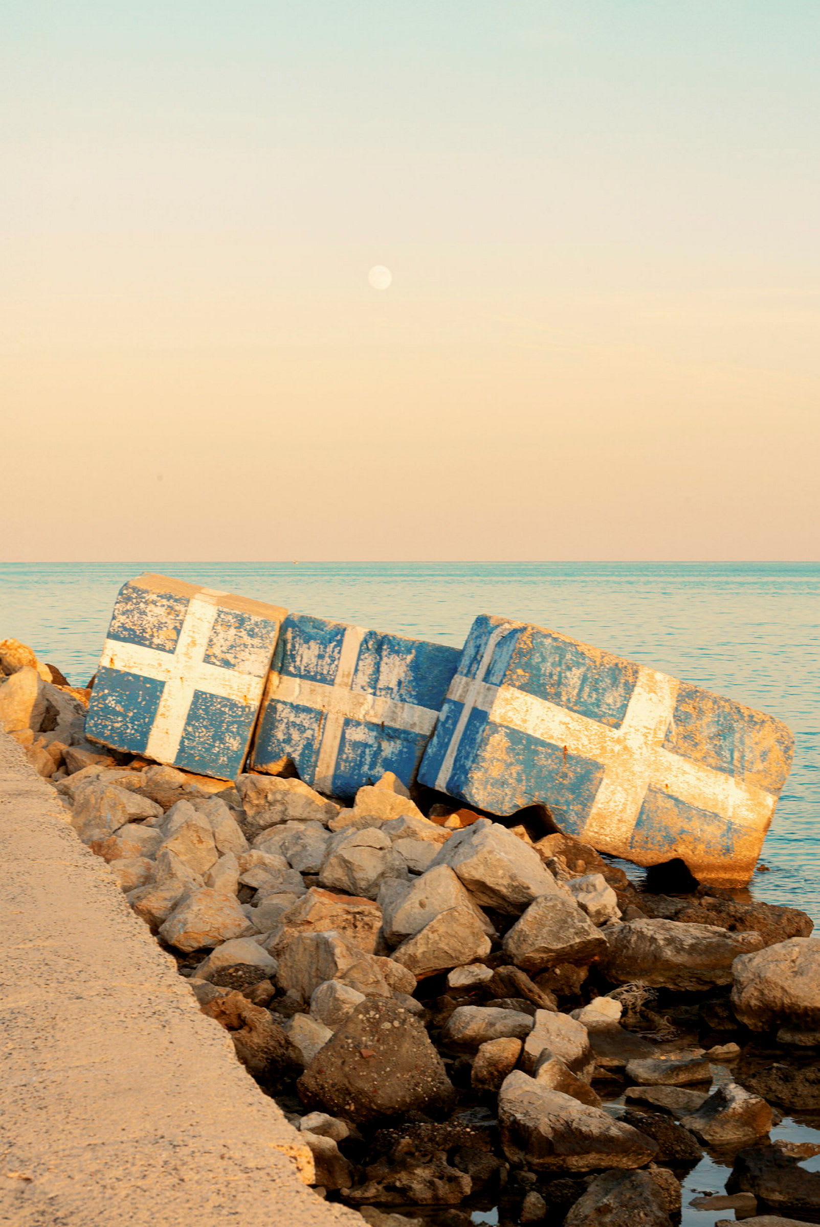 Betongblock målade med vita kors på blå bakgrund vid en stenig strand. (AI-genererad text)
