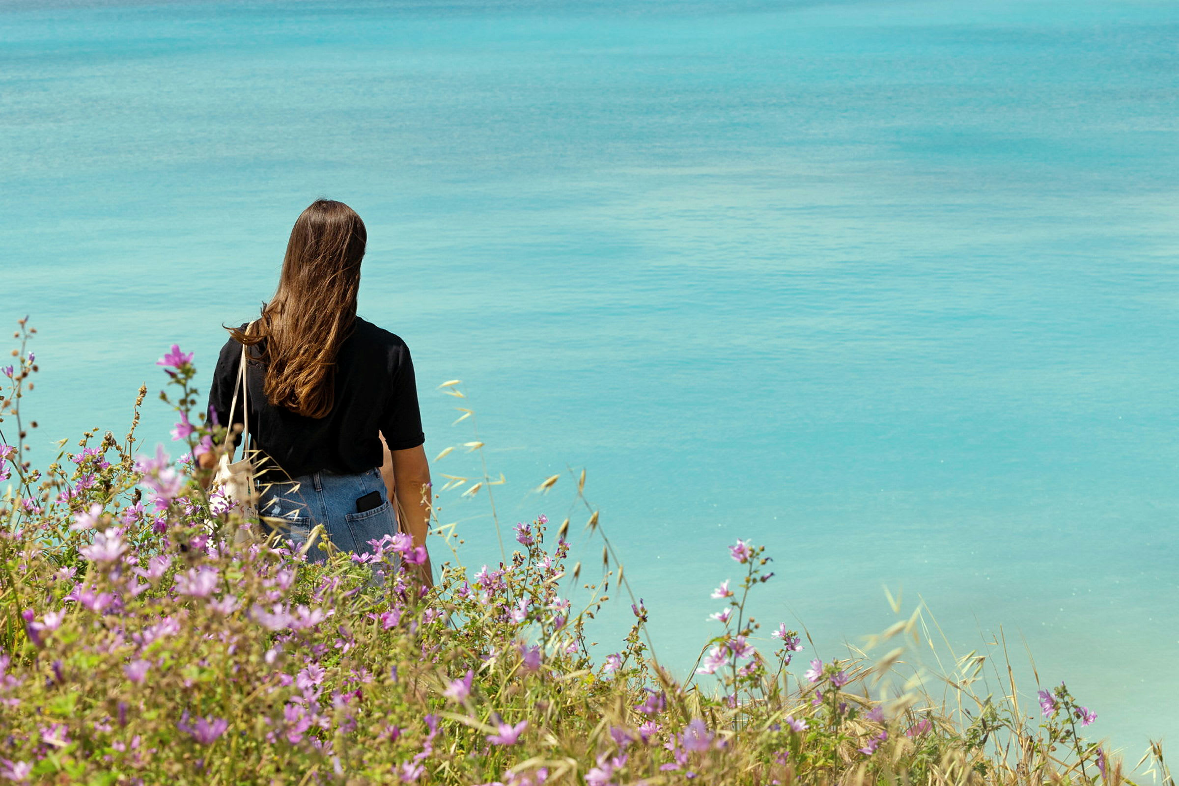 En kvinna står vid kusten med ryggen mot kameran och ser ut mot havet, omgivet av blommande växter. (AI-genererad text)