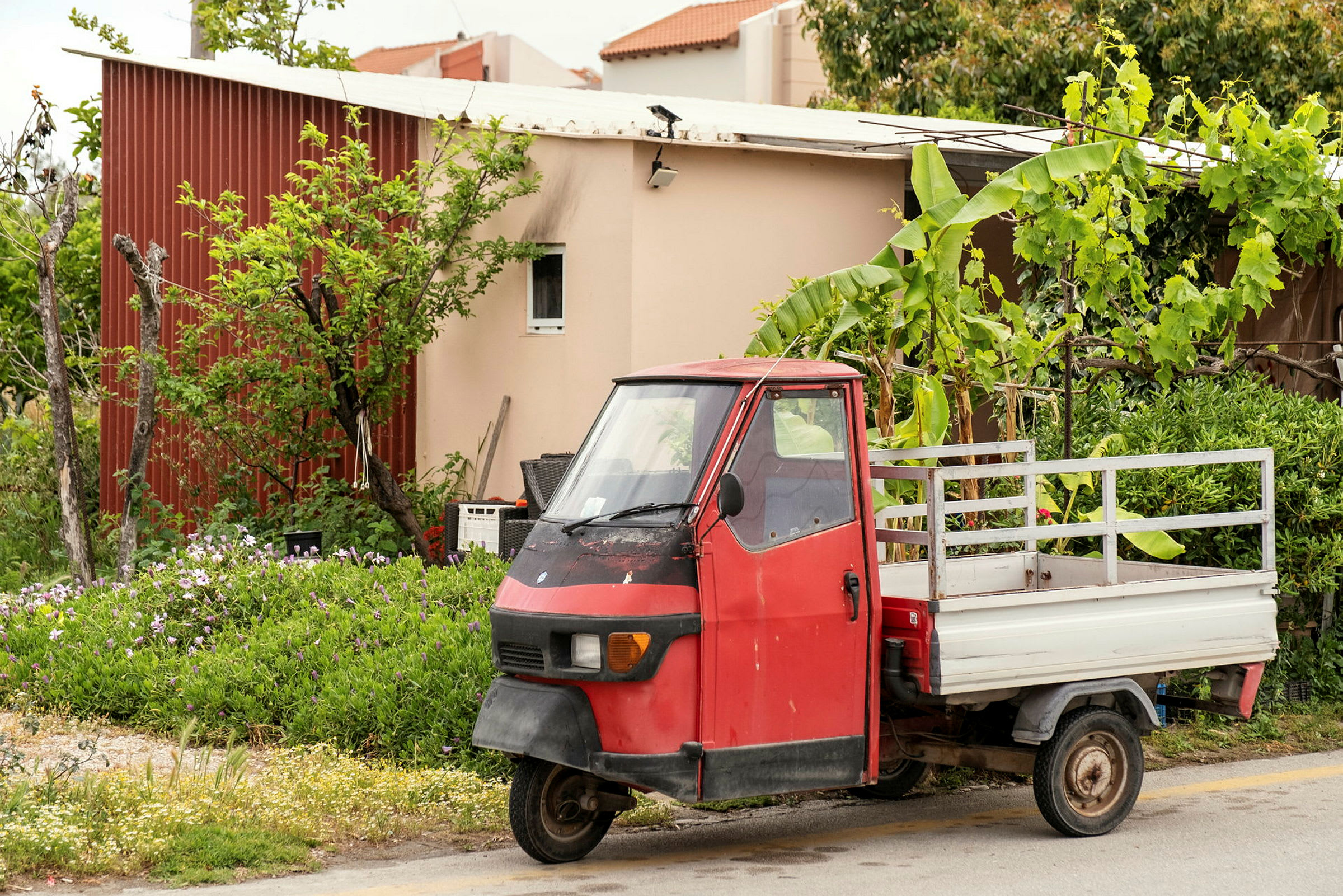 En röd trehjuling lastbil står parkerad framför ett hus med träd och växter runt omkring. (AI-genererad text)