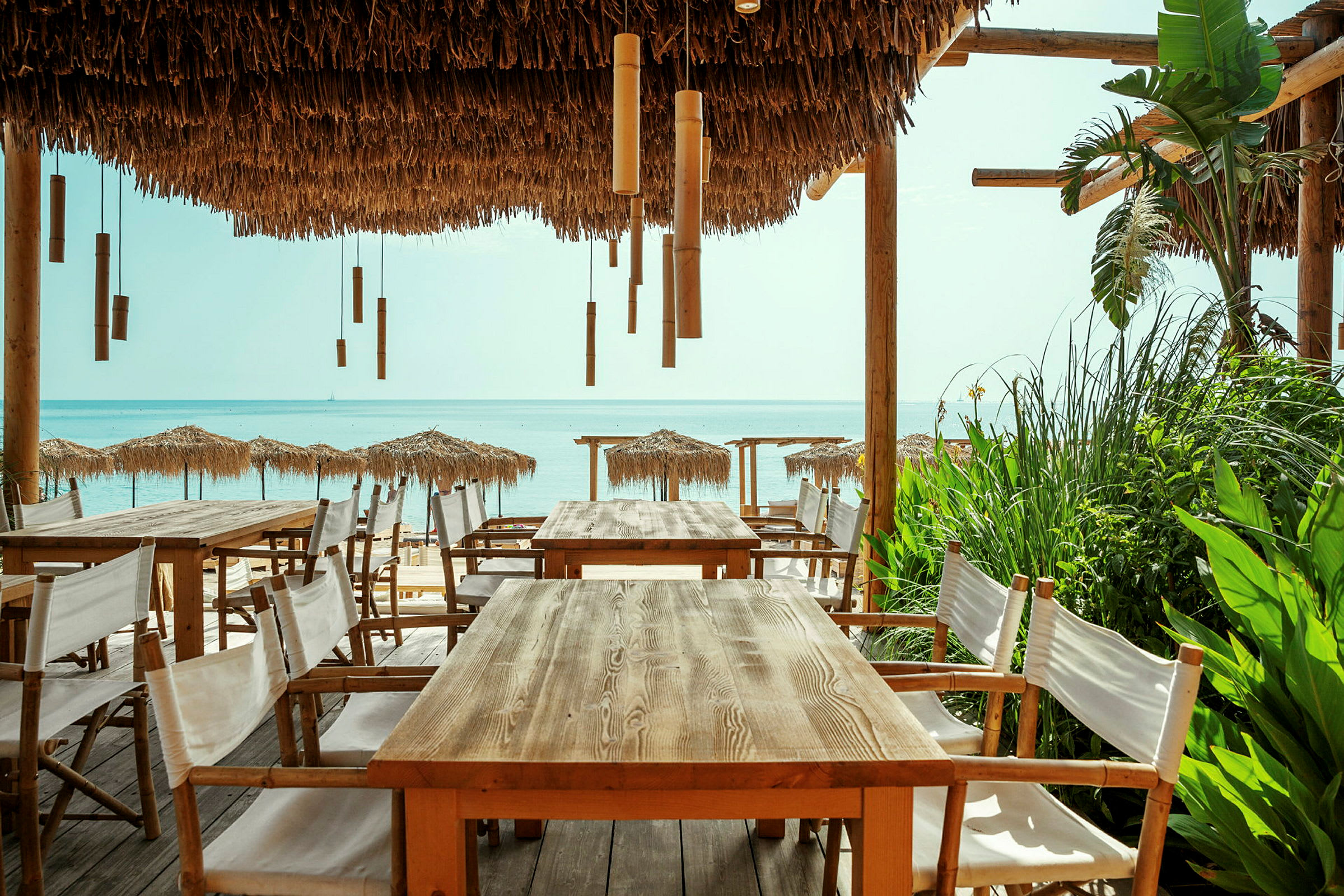 Uteservering med träbord och stolar under en halmtakstäckt pergola, med utsikt över stranden och havet. (AI-genererad text)