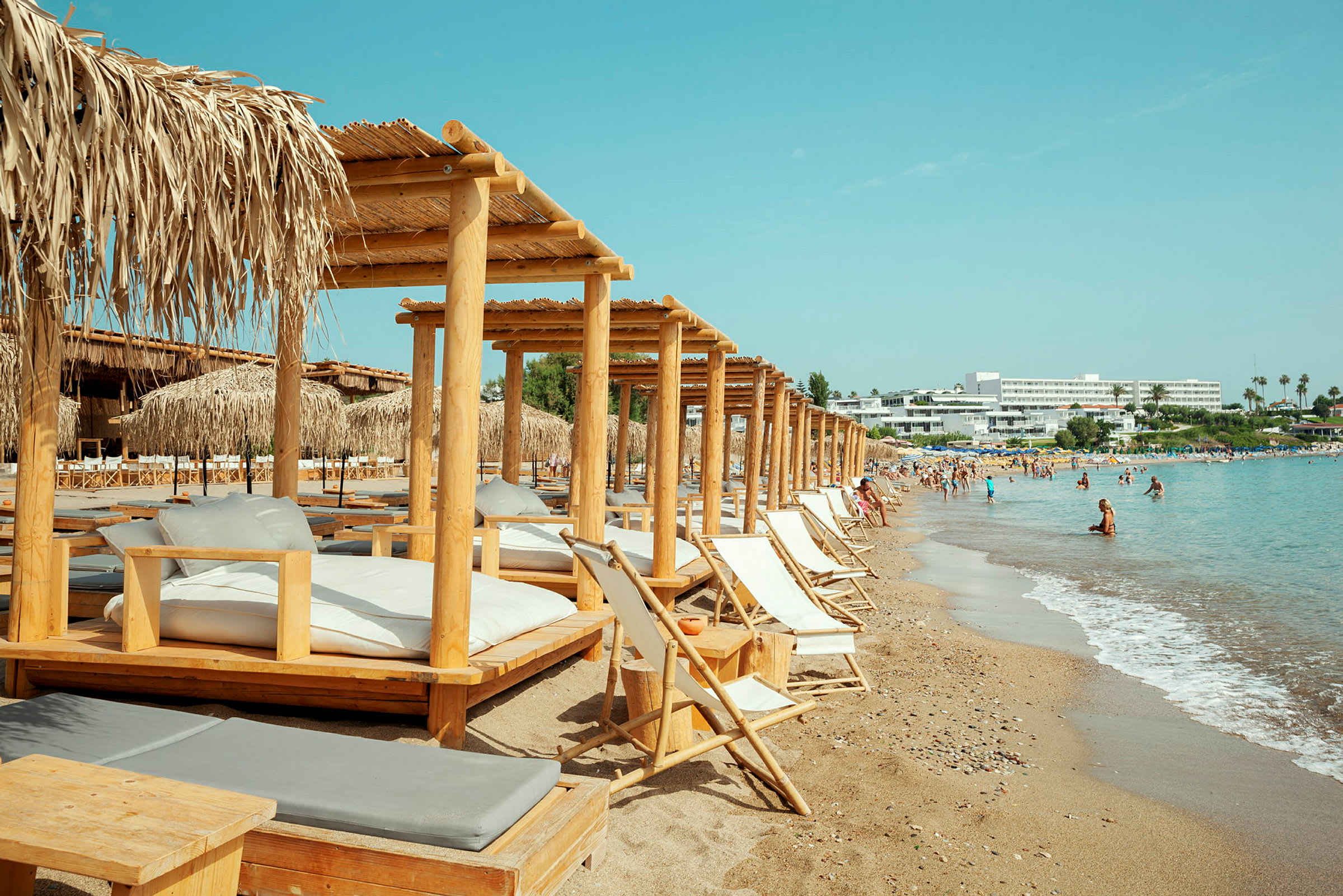 Solstolar och bambusängar på en strand med turkost hav och hotell i bakgrunden. (AI-genererad text)