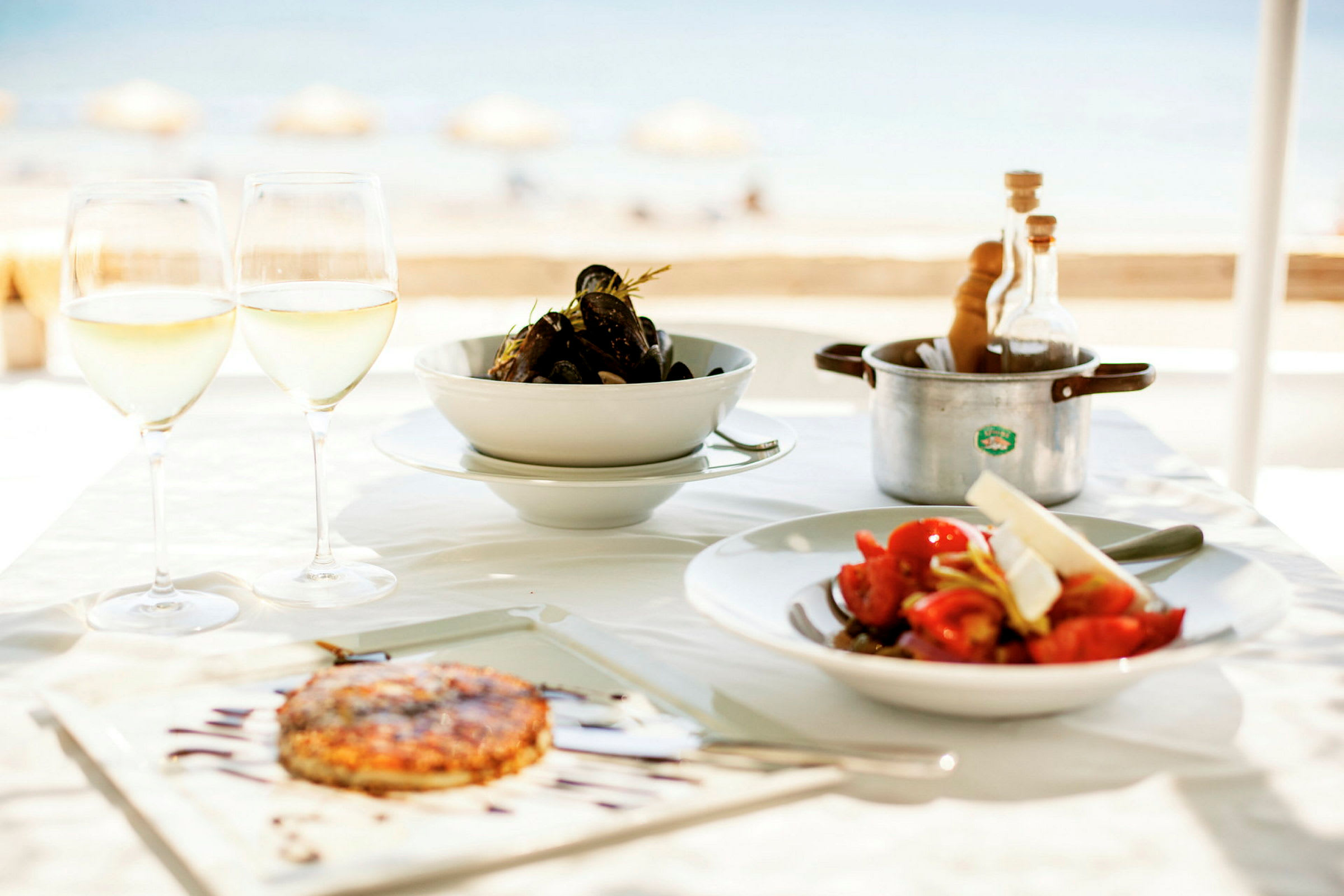 En tallrik med mat, två glas vitt vin och en kastrull på ett dukat bord vid stranden. (AI-genererad text)