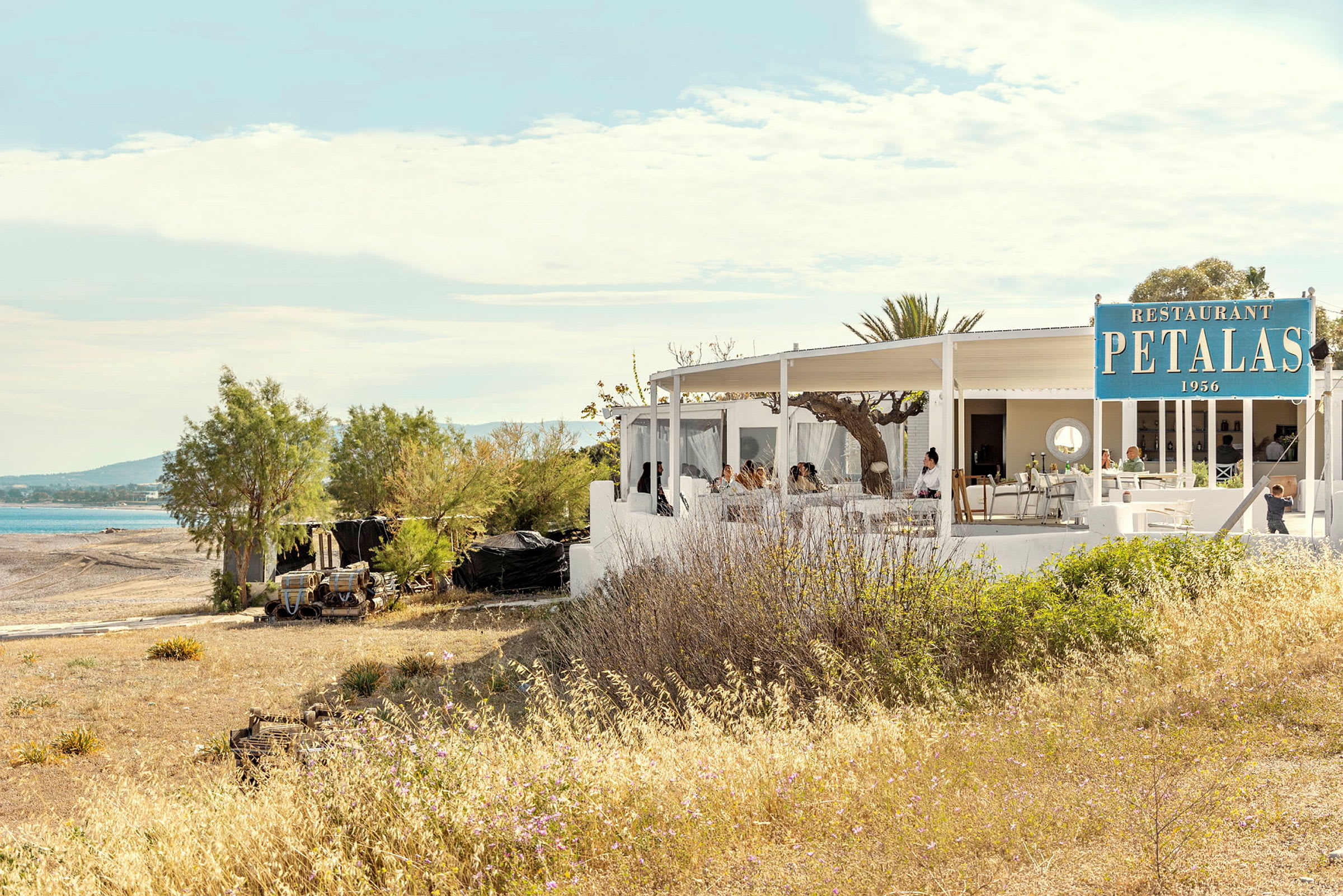 En restaurang med vit byggnad nära en strand med uteservering under tak. (AI-genererad text)