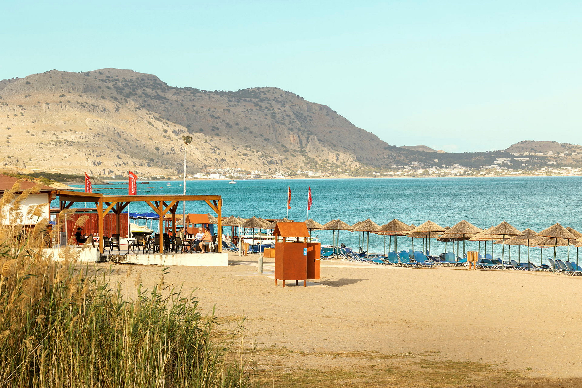 Sandstrand med solstolar och parasoller, samt en restaurang med utsikt över havet och berg i bakgrunden. (AI-genererad text)