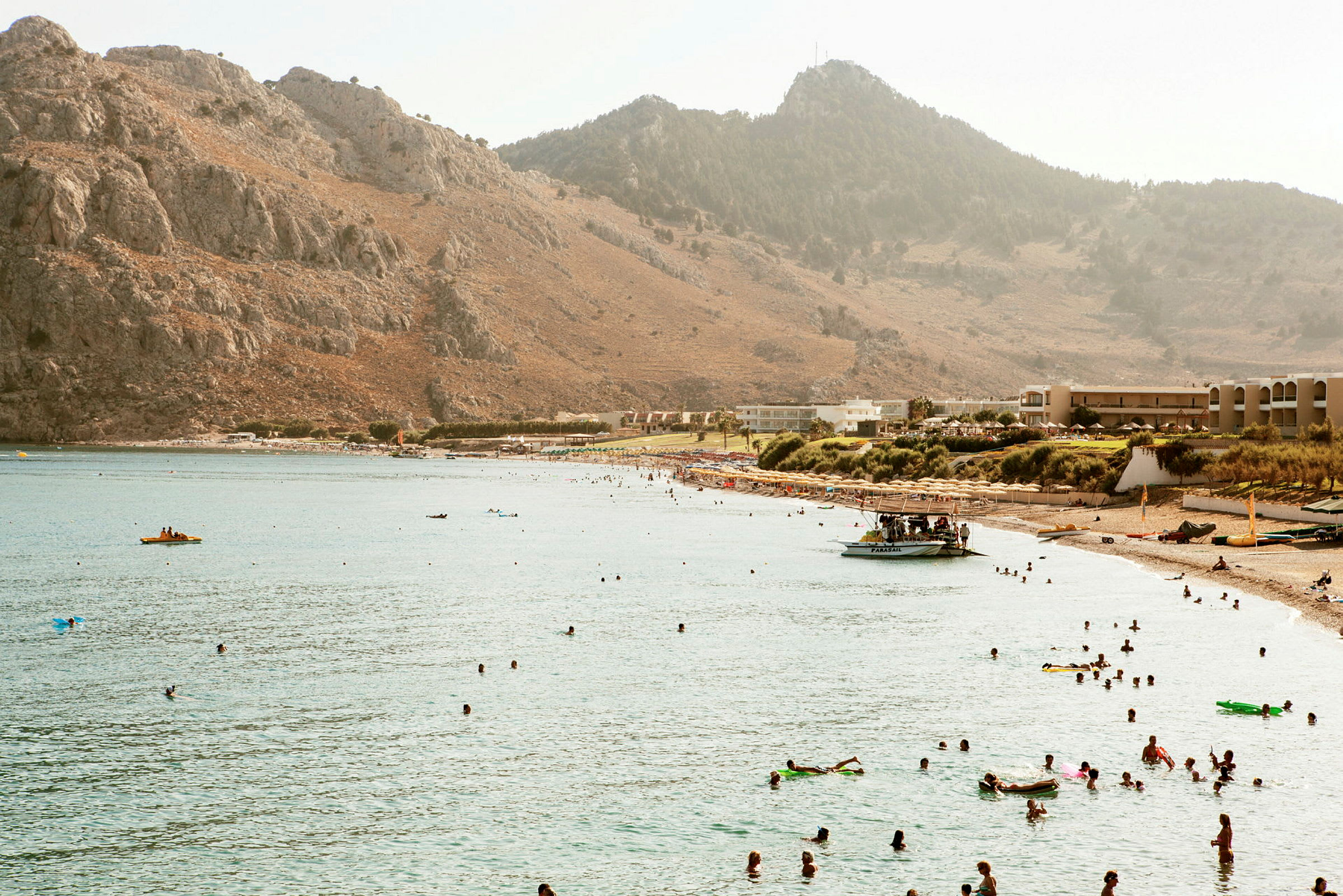 En strand vid en vik omgiven av berg, med människor som badar i vattnet och solbadar på stranden. (AI-genererad text)