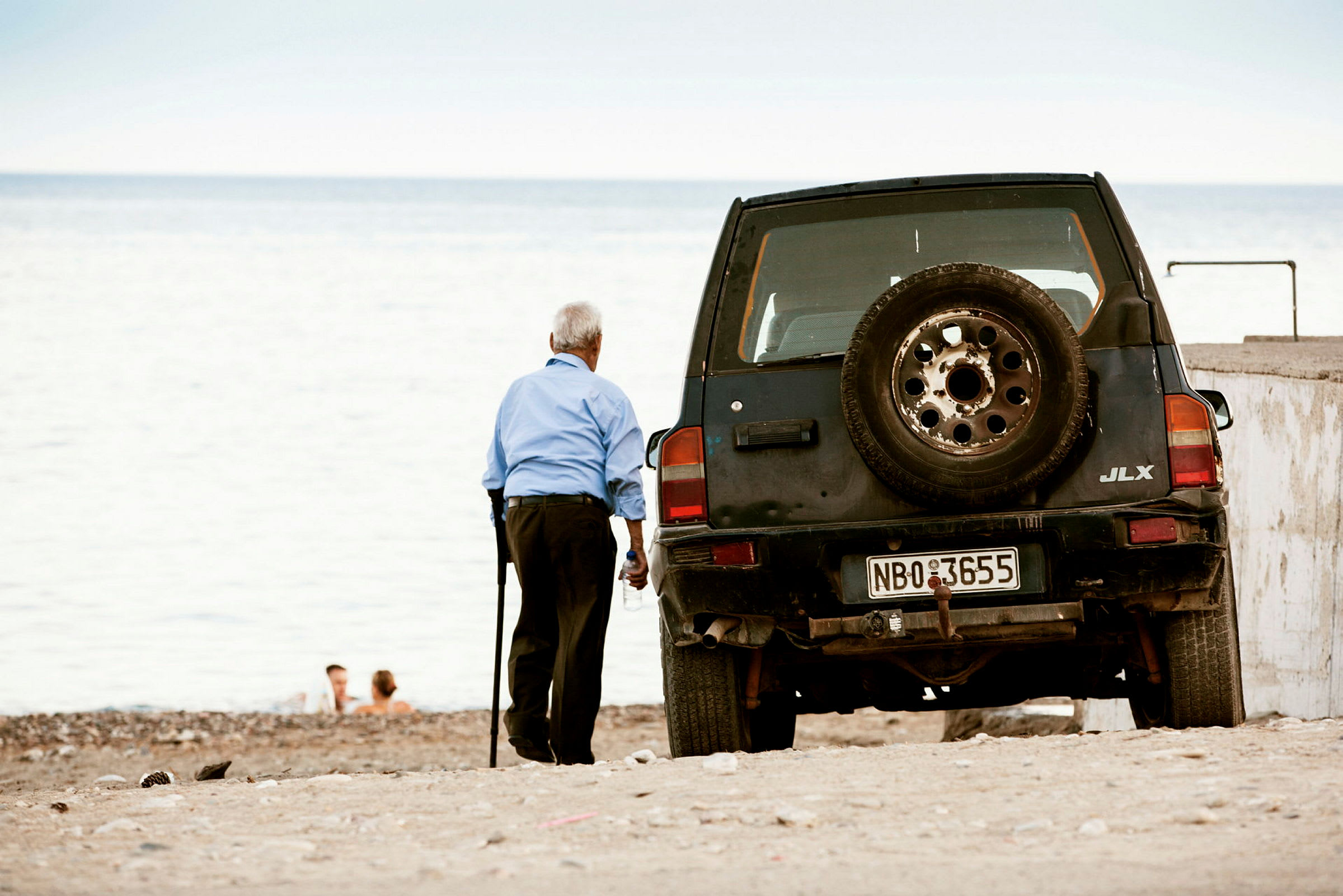 En äldre person står på en strand bredvid en svart bil med reservdäck på baksidan. (AI-genererad text)