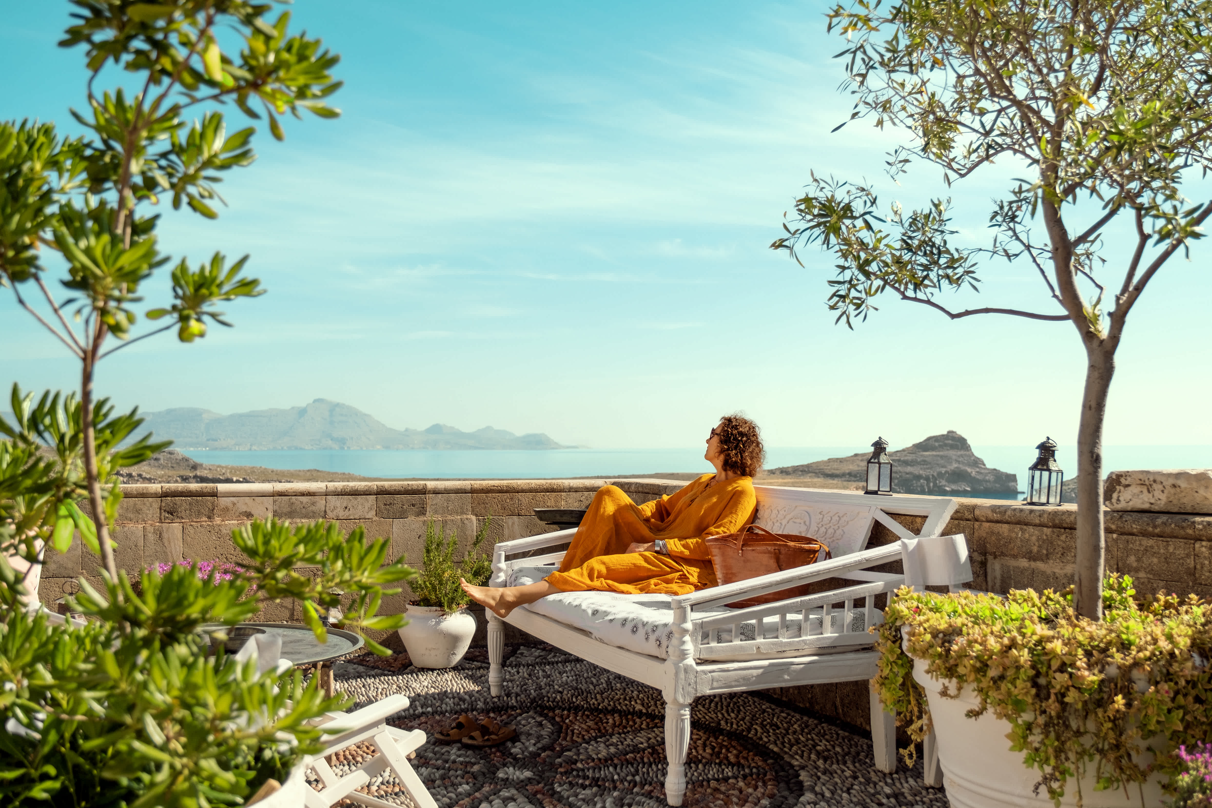 En person sitter avslappnad på en vit bänk på en terrass, omgiven av grönska med utsikt över havet och bergen. (AI-genererad text)