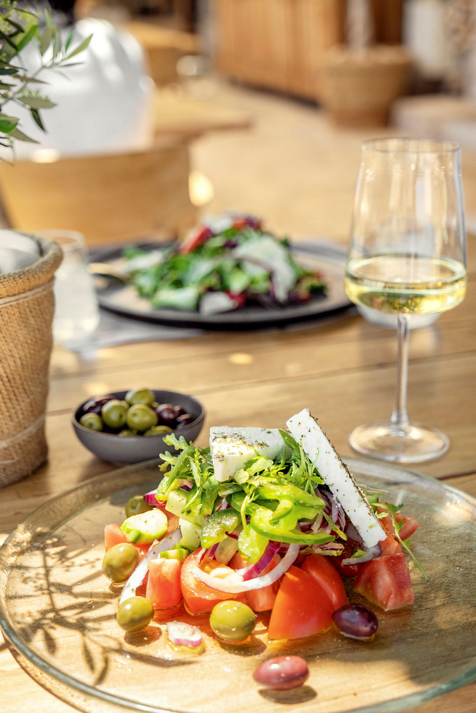 Bild på en tallrik med sallad, fetaost och oliver samt ett glas vitt vin på ett träbord. (AI-genererad text)