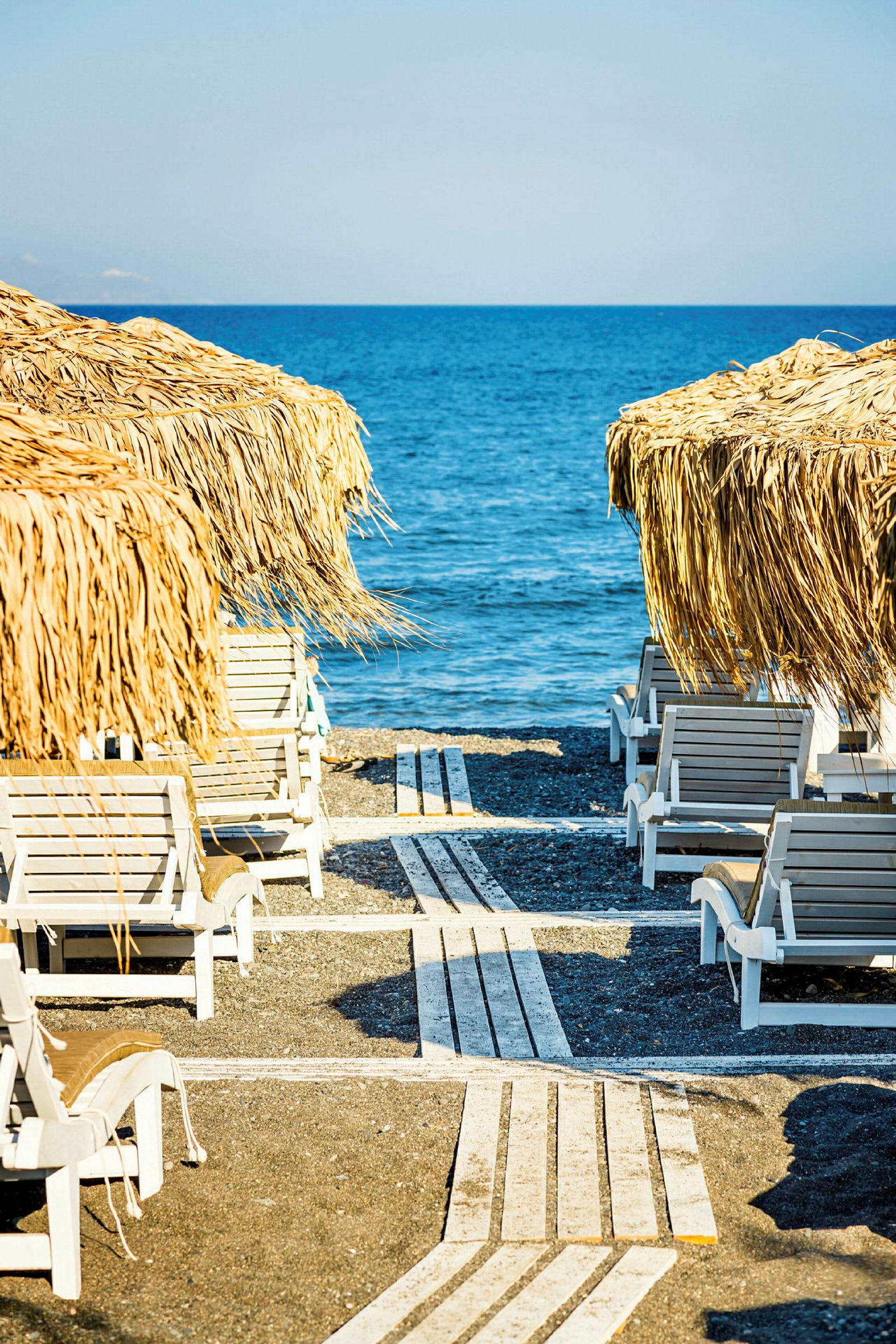 Solstolar på en strand med parasoller av växtmaterial och havet i bakgrunden. (AI-genererad text)