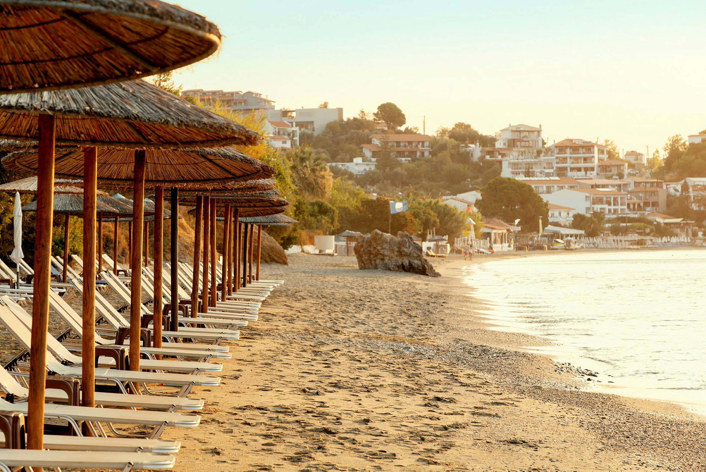 Solstolar och parasoller på en strand med hav och byggnader i bakgrunden. (AI-genererad text)