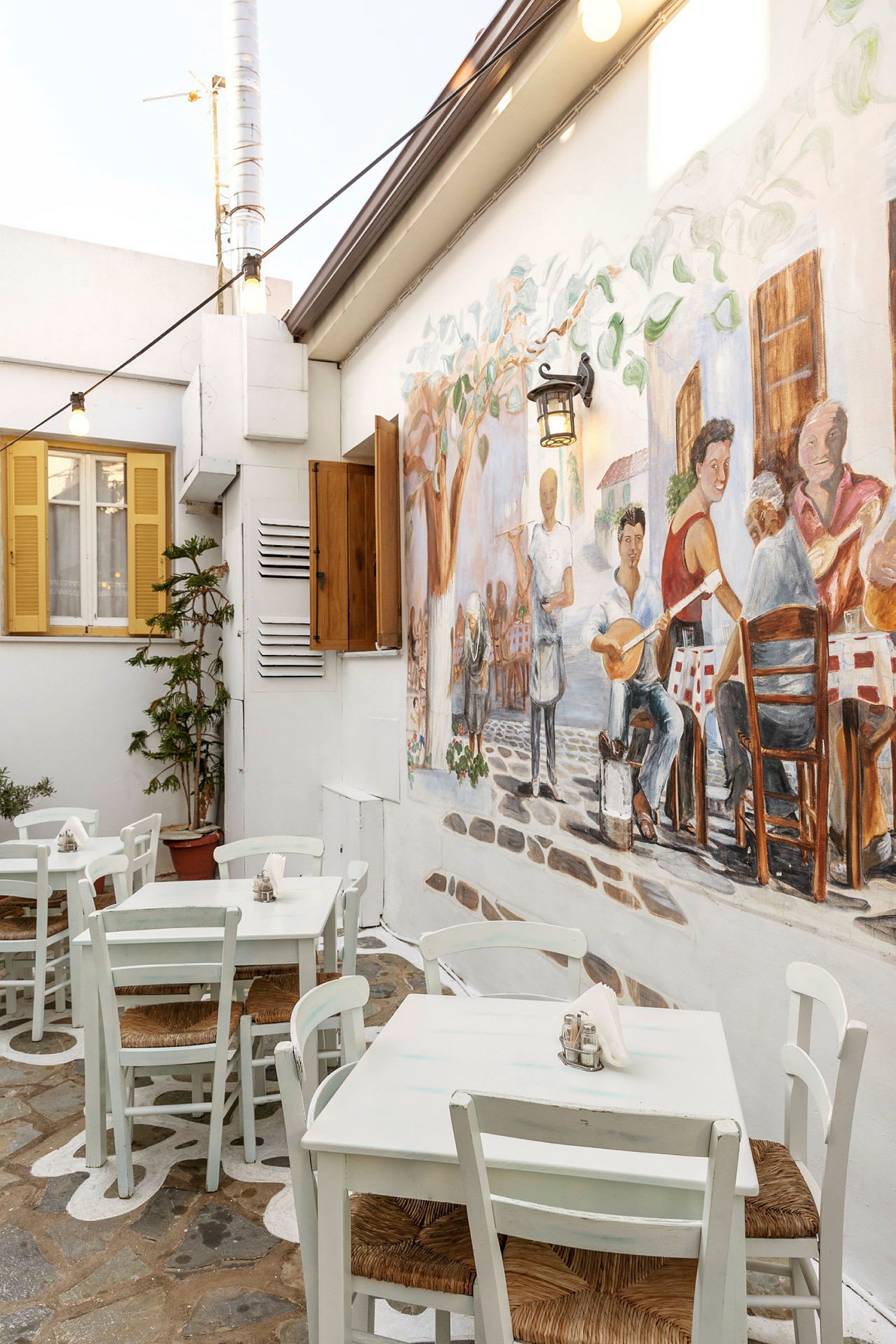 Utomhusområde med vita bord och stolar, vägg dekorerad med målning av människor i en restaurangmiljö. (AI-genererad text)