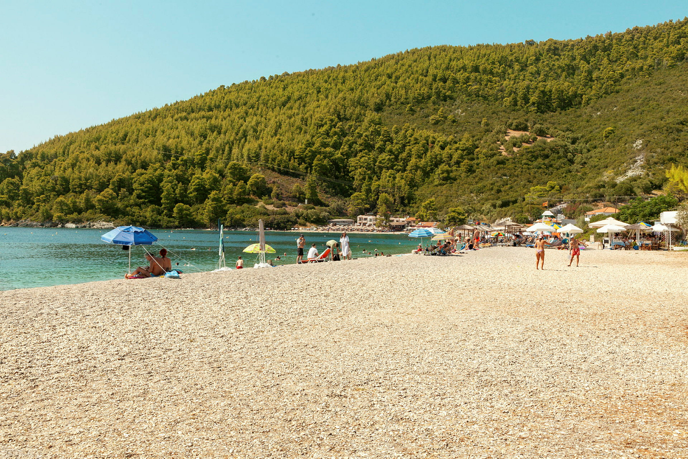 En strand med solbadande människor, parasoller och omgivande gröna kullar. (AI-genererad text)
