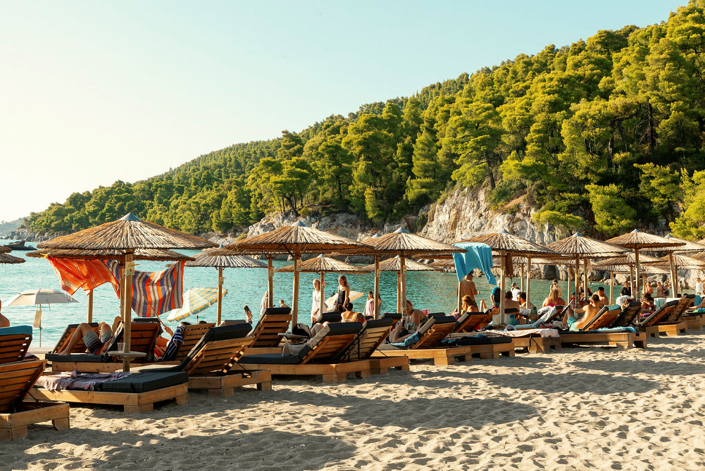 Personer som kopplar av på en strand med solsängar och parasoller, med en grönskande bergssluttning i bakgrunden. (AI-genererad text)