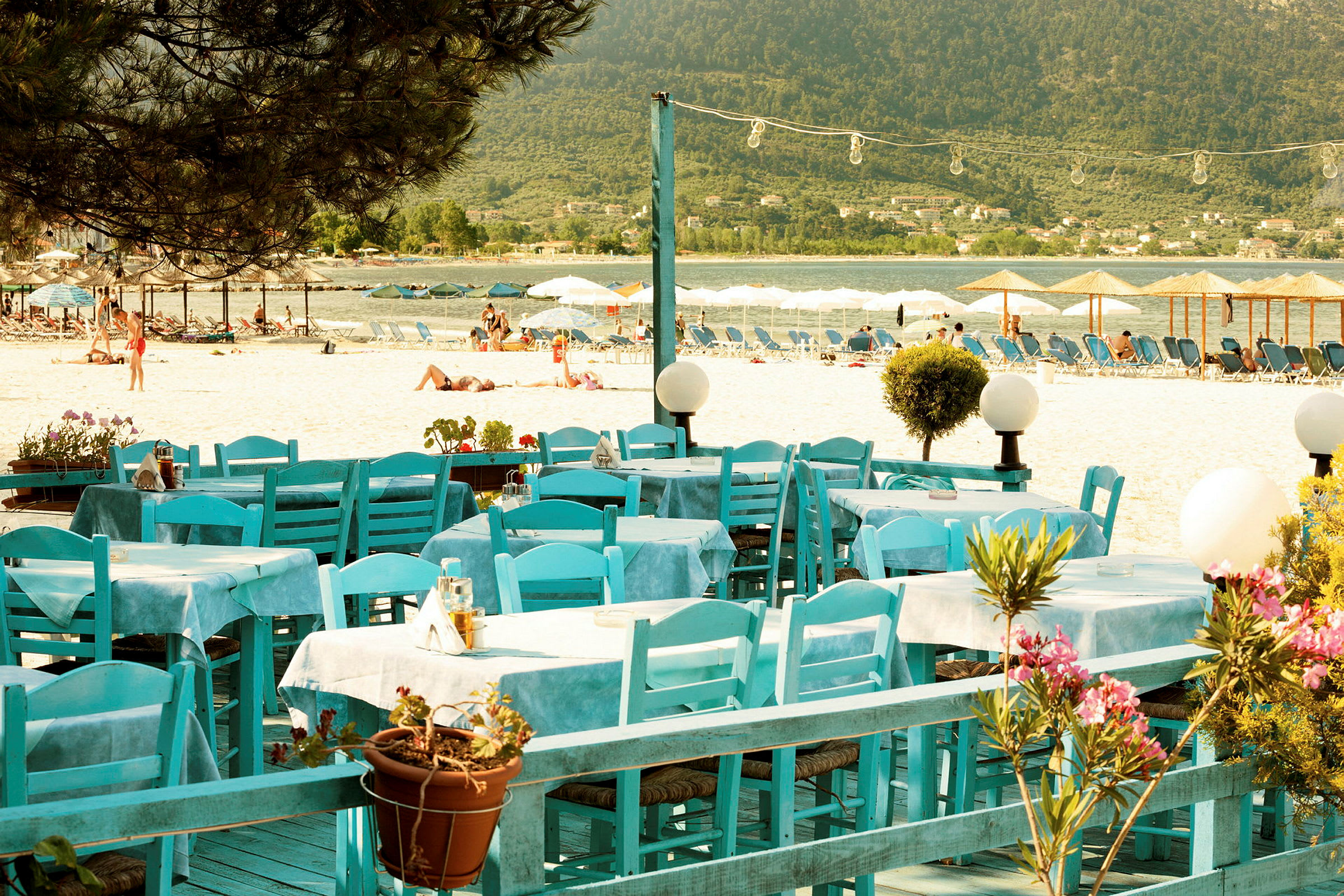 Restaurangbord med blå stolar vid en sandstrand med solstolar och parasoller i bakgrunden. (AI-genererad text)