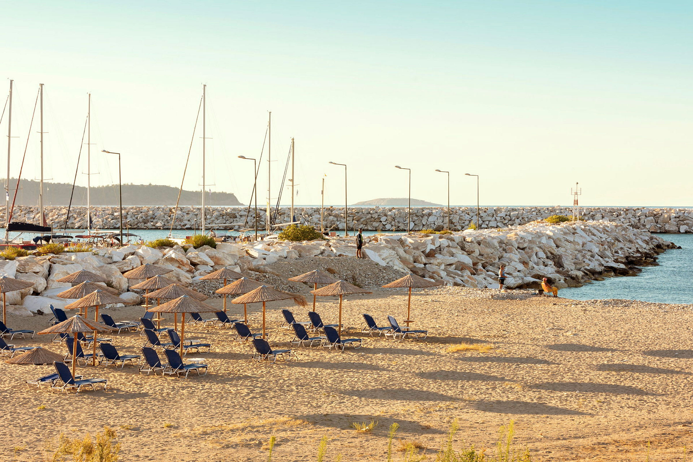 Strand med solstolar och parasoller, i bakgrunden syns en hamn med segelbåtar. (AI-genererad text)